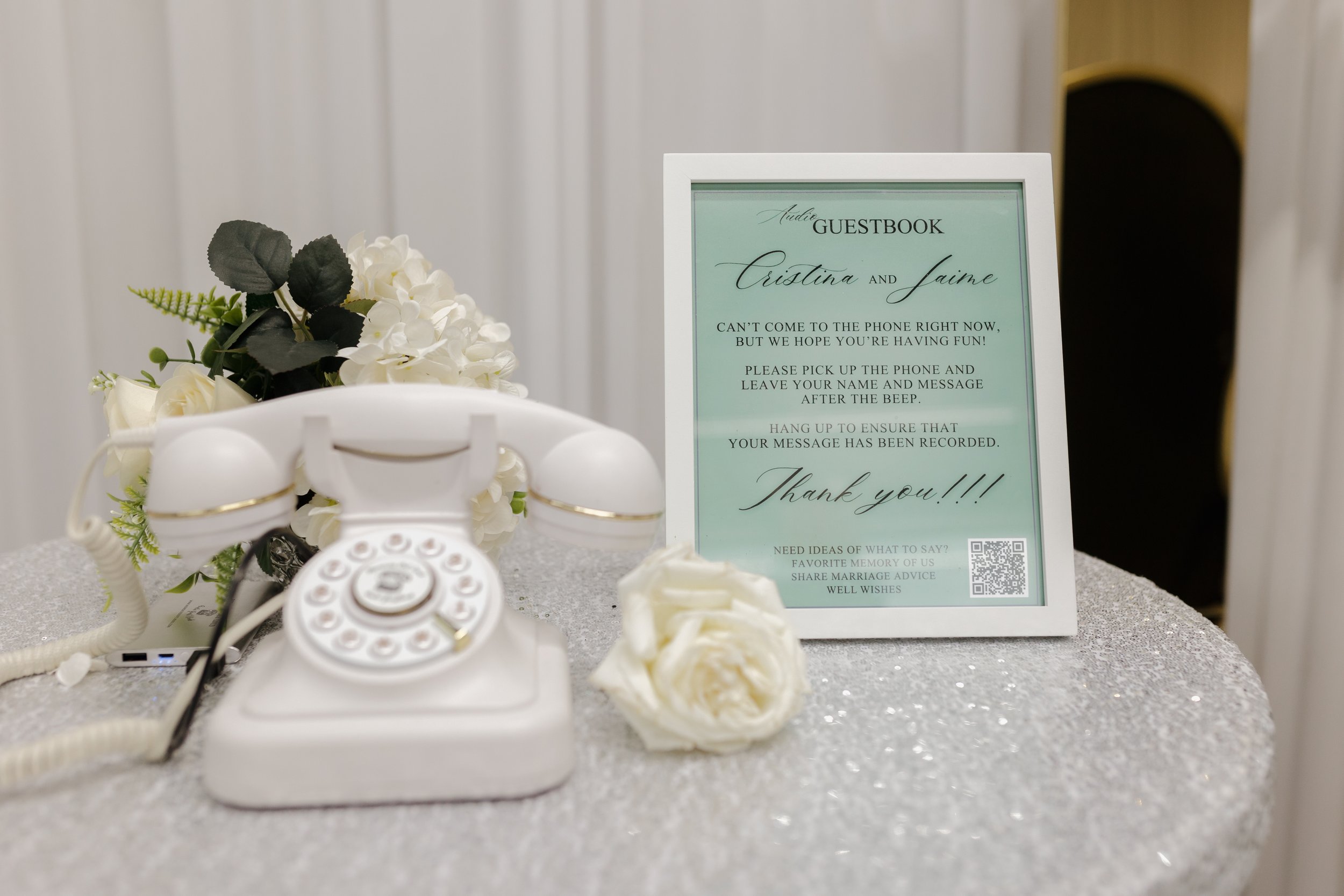 An elegant white vintage audio guestbook phone styled on a shimmering white sequin table alongside white roses and framed mint green wedding signage.