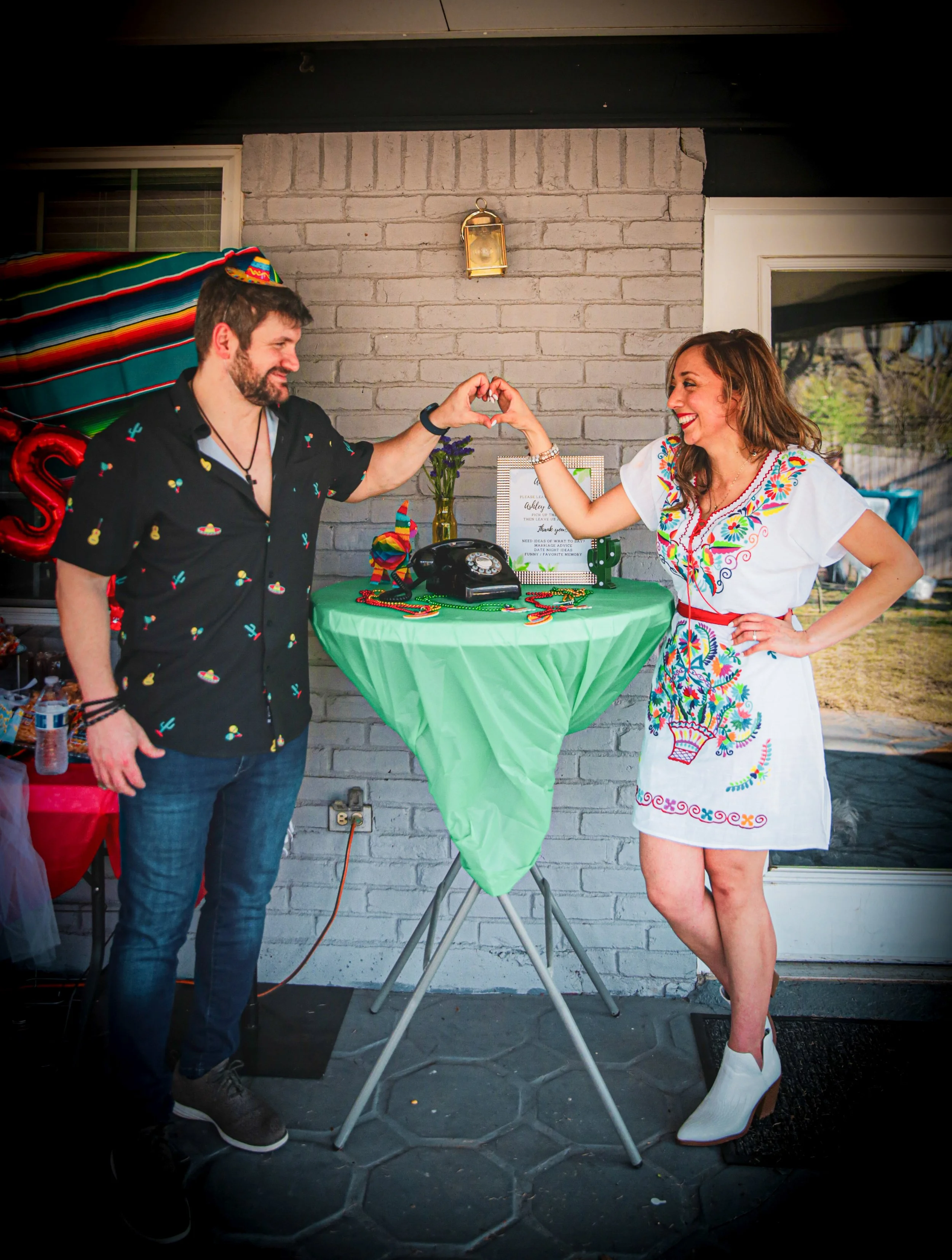 A happy couple dressed in festive fiesta attire making a heart shape with their hands next to a black rotary audio guestbook phone setup at an outdoor fiesta celebration.