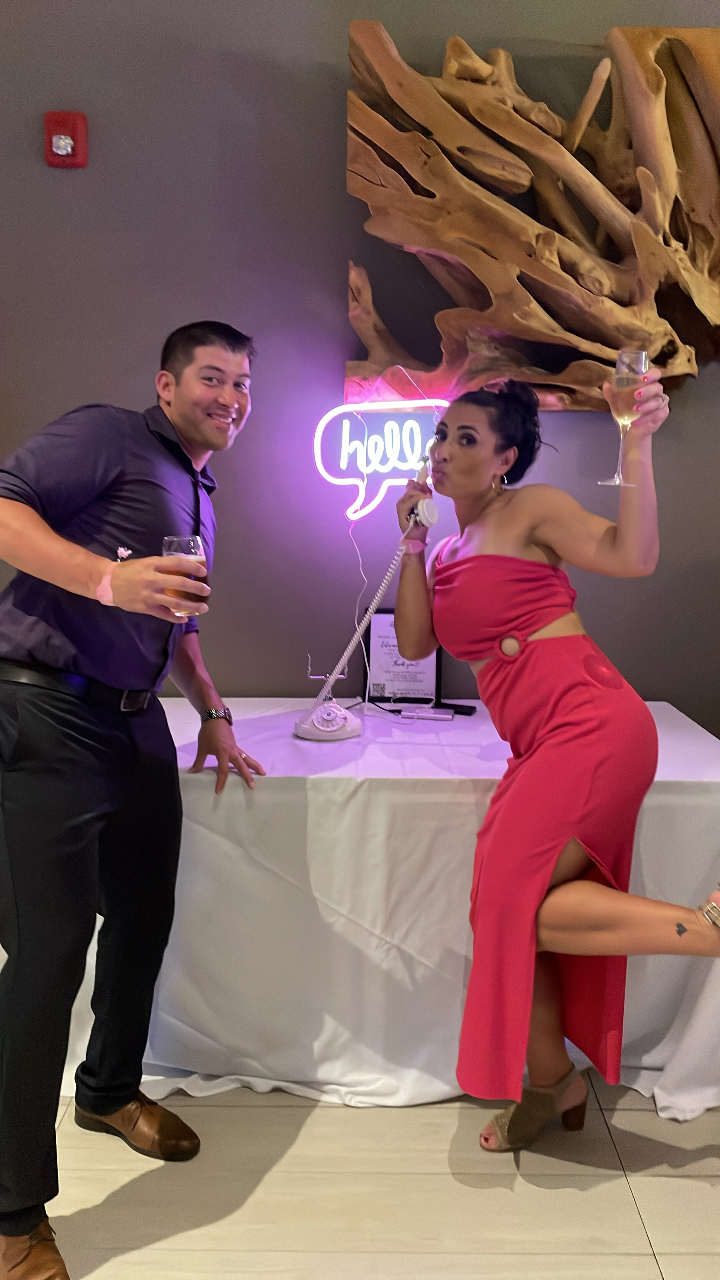 Event guests posing playfully with drinks in hand while using a vintage Memories Out Loud audio guestbook phone setup in front of a glowing pink neon 'hello' sign.