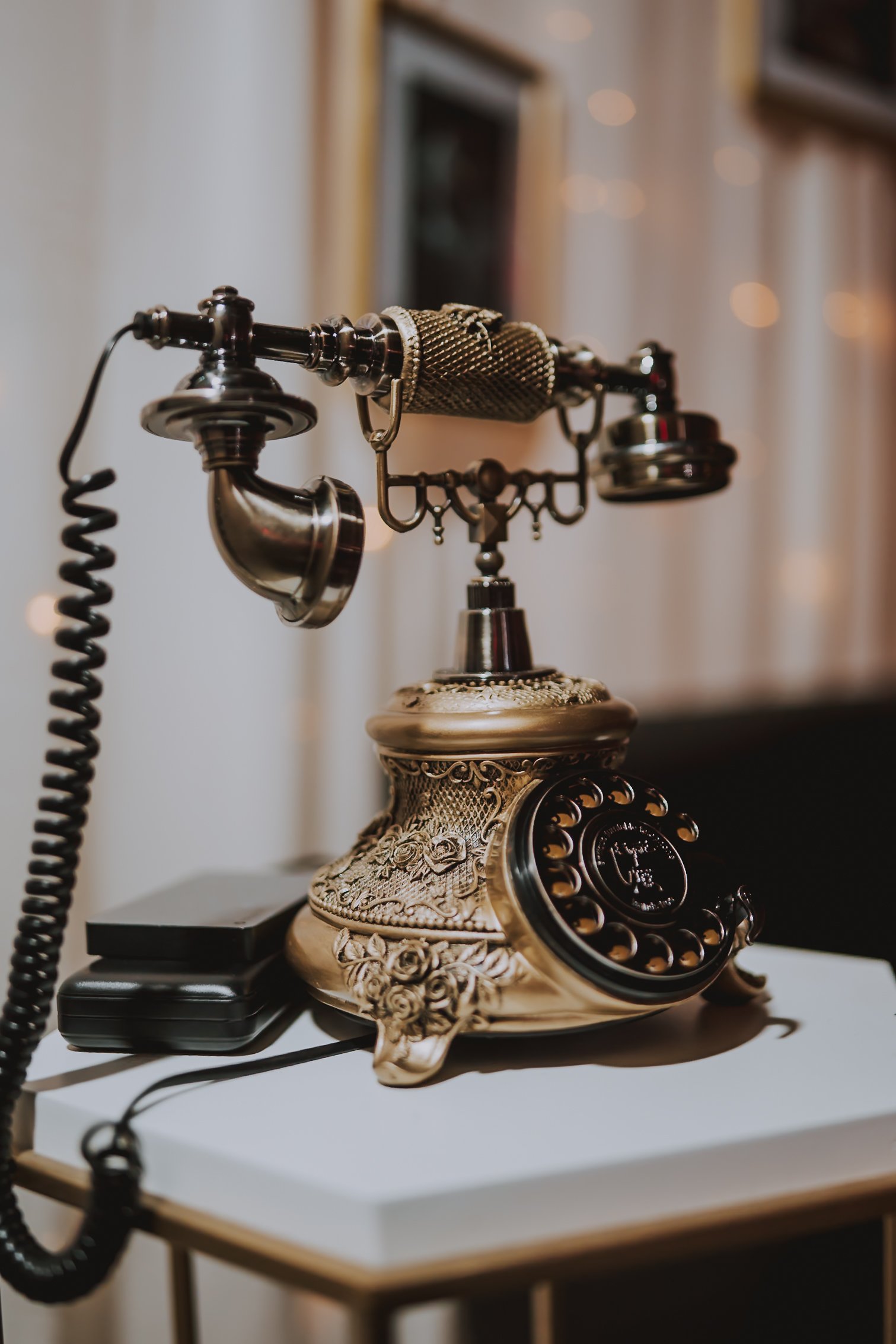 Detailed close-up shot of an ornate antique brass audio guestbook phone by Memories Out Loud displayed on a white pedestal with warm background lighting.