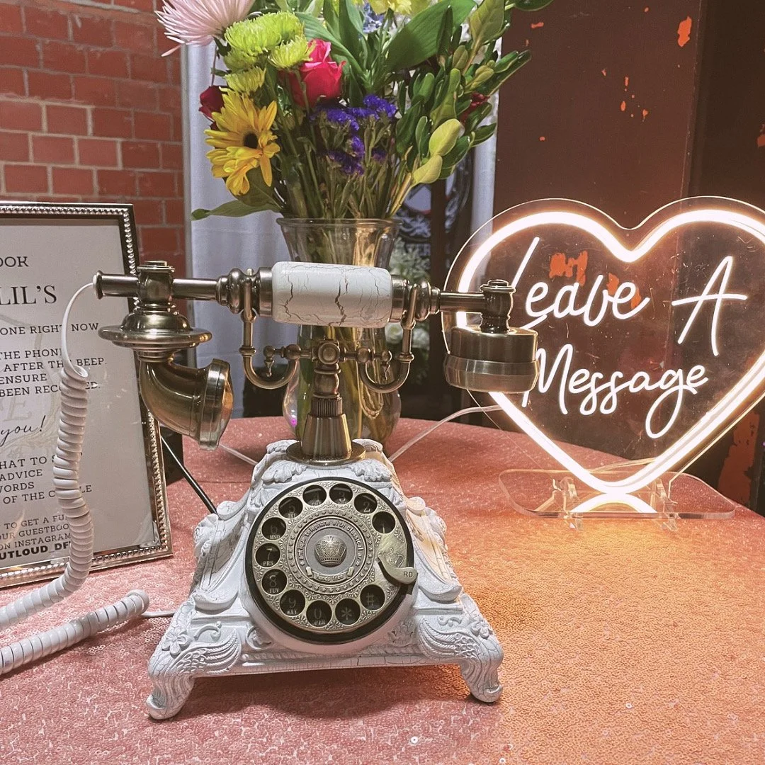 Ornate Charlotte white and brass vintage audio guestbook phone styled on a peach sequin table next to a glowing heart-shaped 'Leave A Message' neon sign and colorful floral centerpiece.