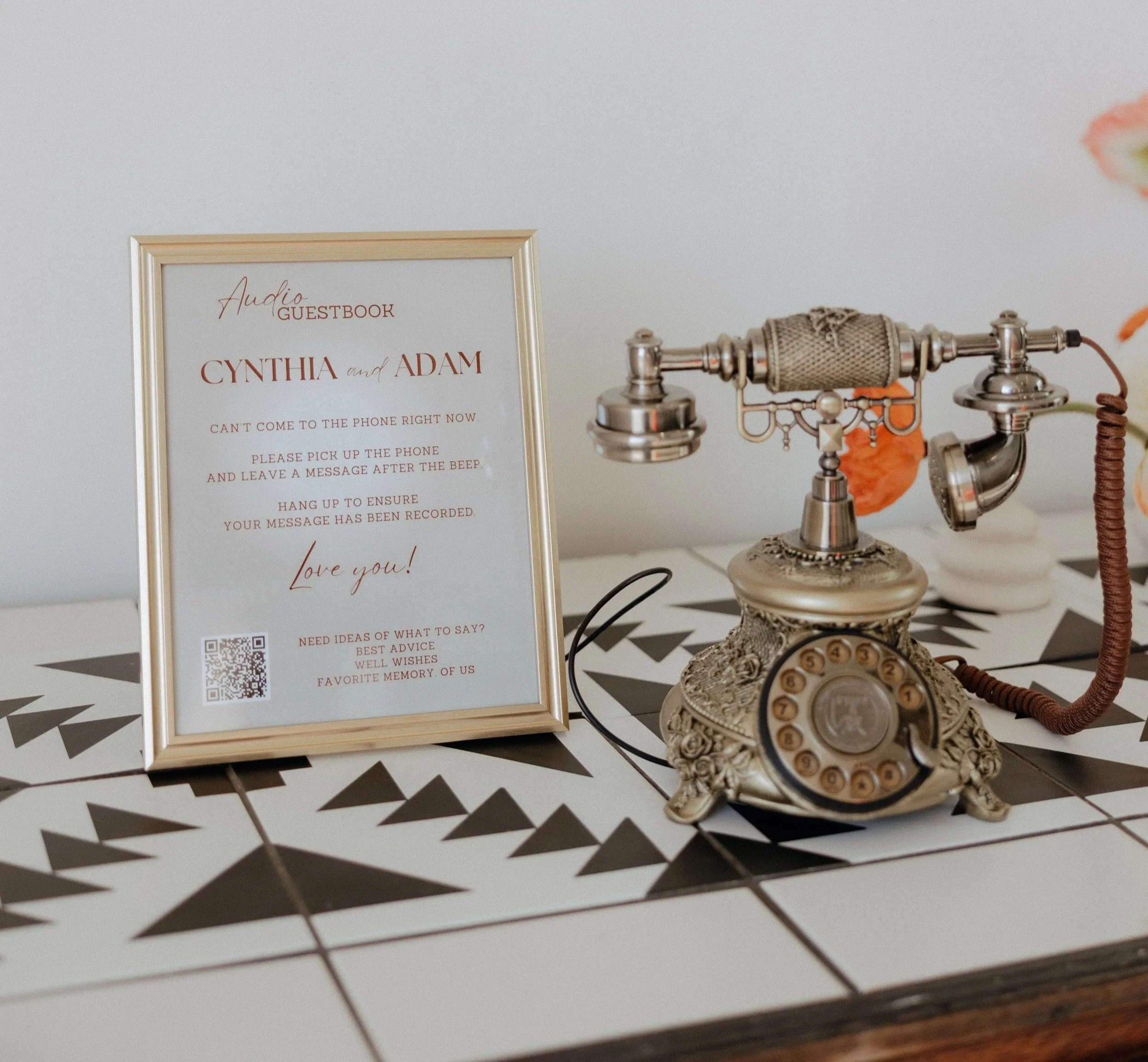 An antique brass audio guestbook phone next to a framed 'Audio Guestbook Cynthia and Adam' instruction sign on a geometric patterned table.