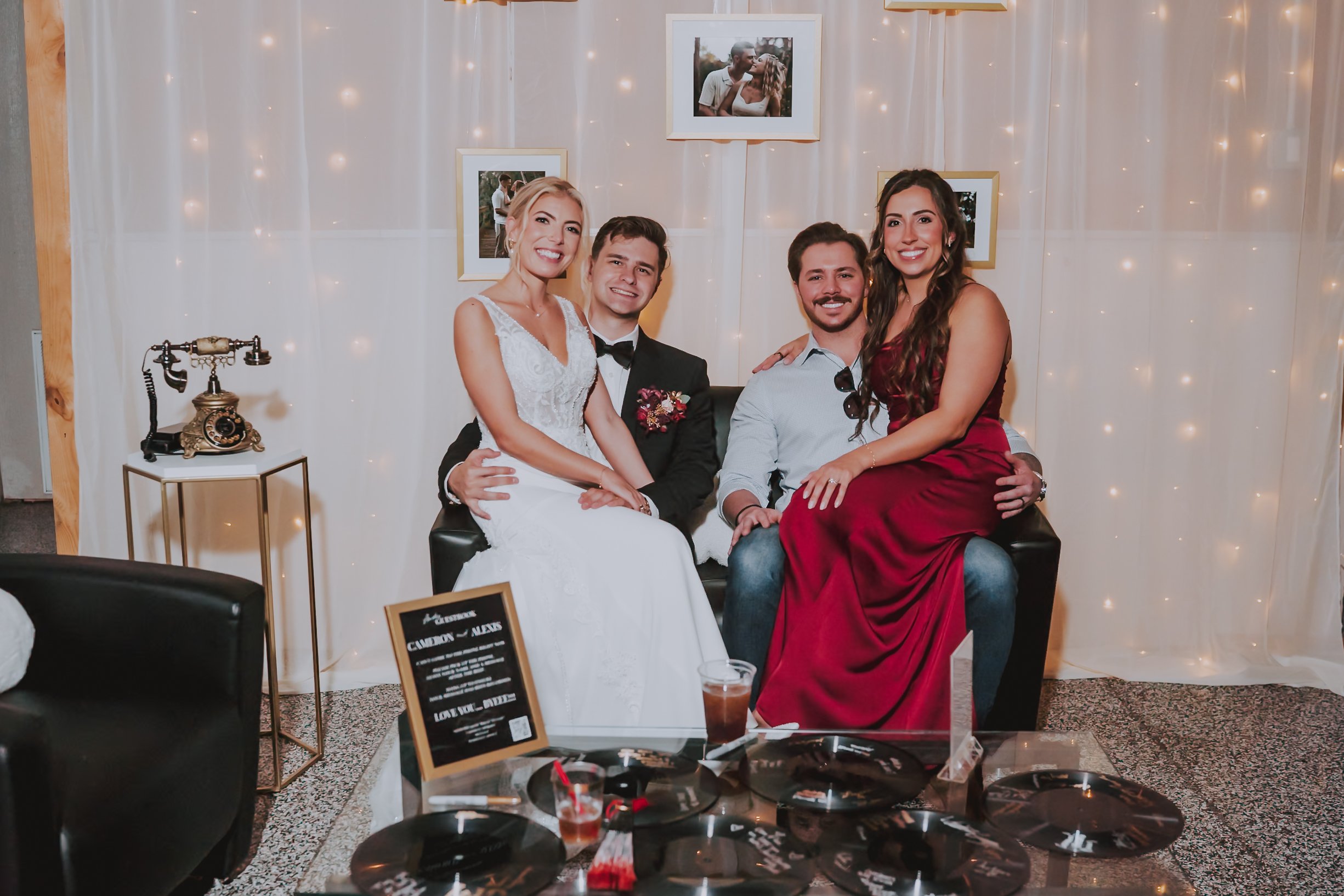 Bride, groom, and wedding guests sitting together in a cozy reception lounge area next to a dark antique audio guestbook phone set up on a gold side table.