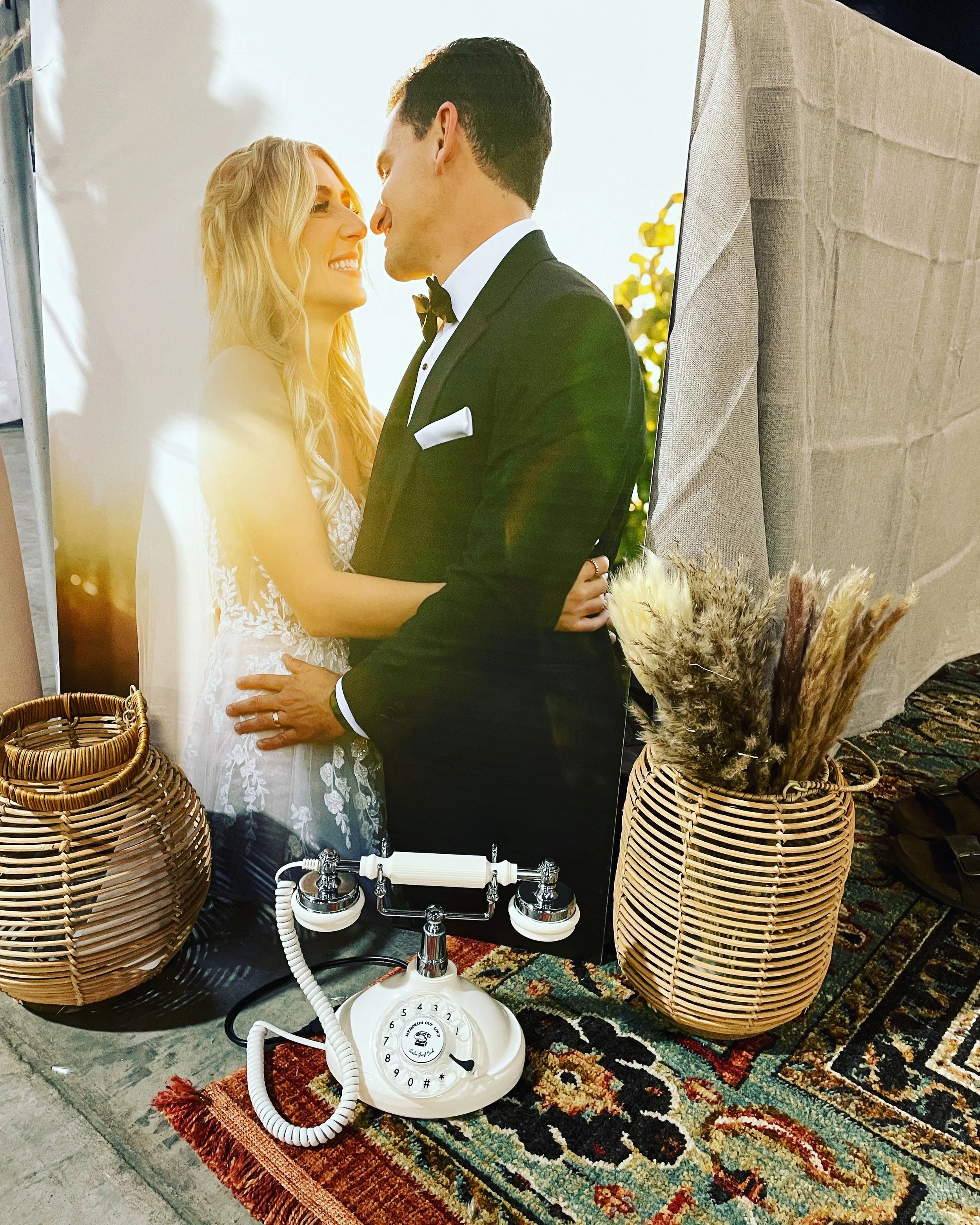 A cream and chrome Memories Out Loud vintage audio guestbook phone set on a vintage oriental rug surrounded by woven lanterns, pampas grass, and a bride and groom portrait backdrop.