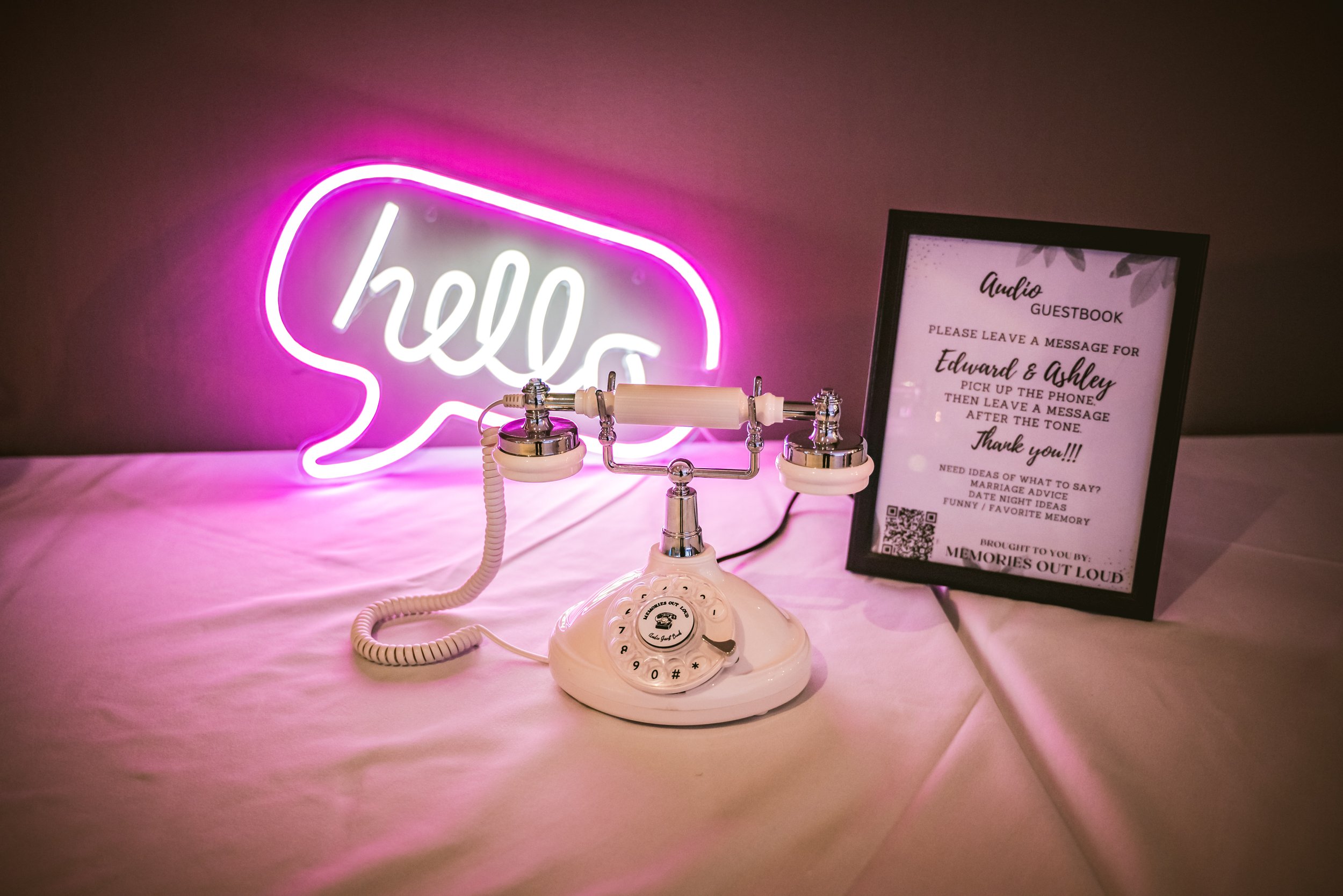 A white and chrome vintage audio guestbook phone set up on a table next to a glowing pink speech-bubble 'hello' neon sign and framed event instructions.