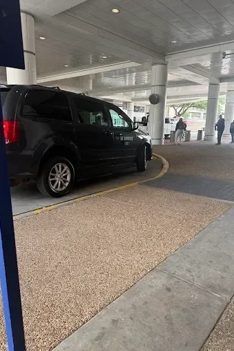 A black minivan parked in a designated area outside a building, with a curved sidewalk and several people in the background.