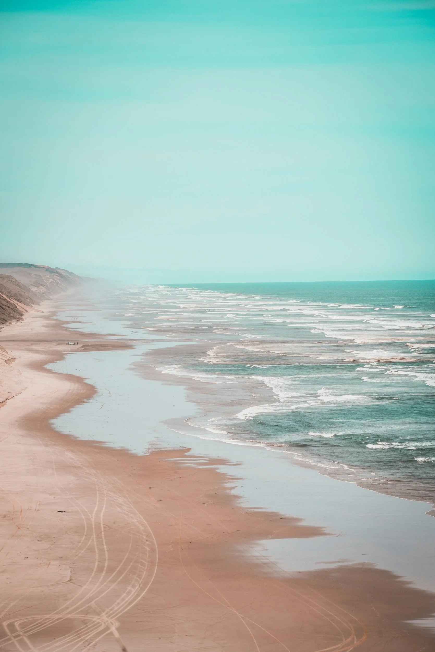 A beach with sand, ocean waves, and cliffs in the background under a clear sky.