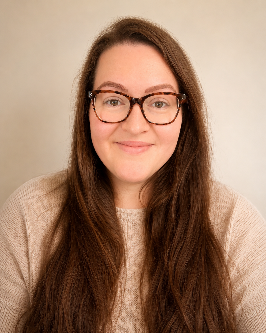 Front portrait of a young woman with long brown hair, glasses with a tortoise shell frame, and a beige sweater, smiling softly against a plain background.