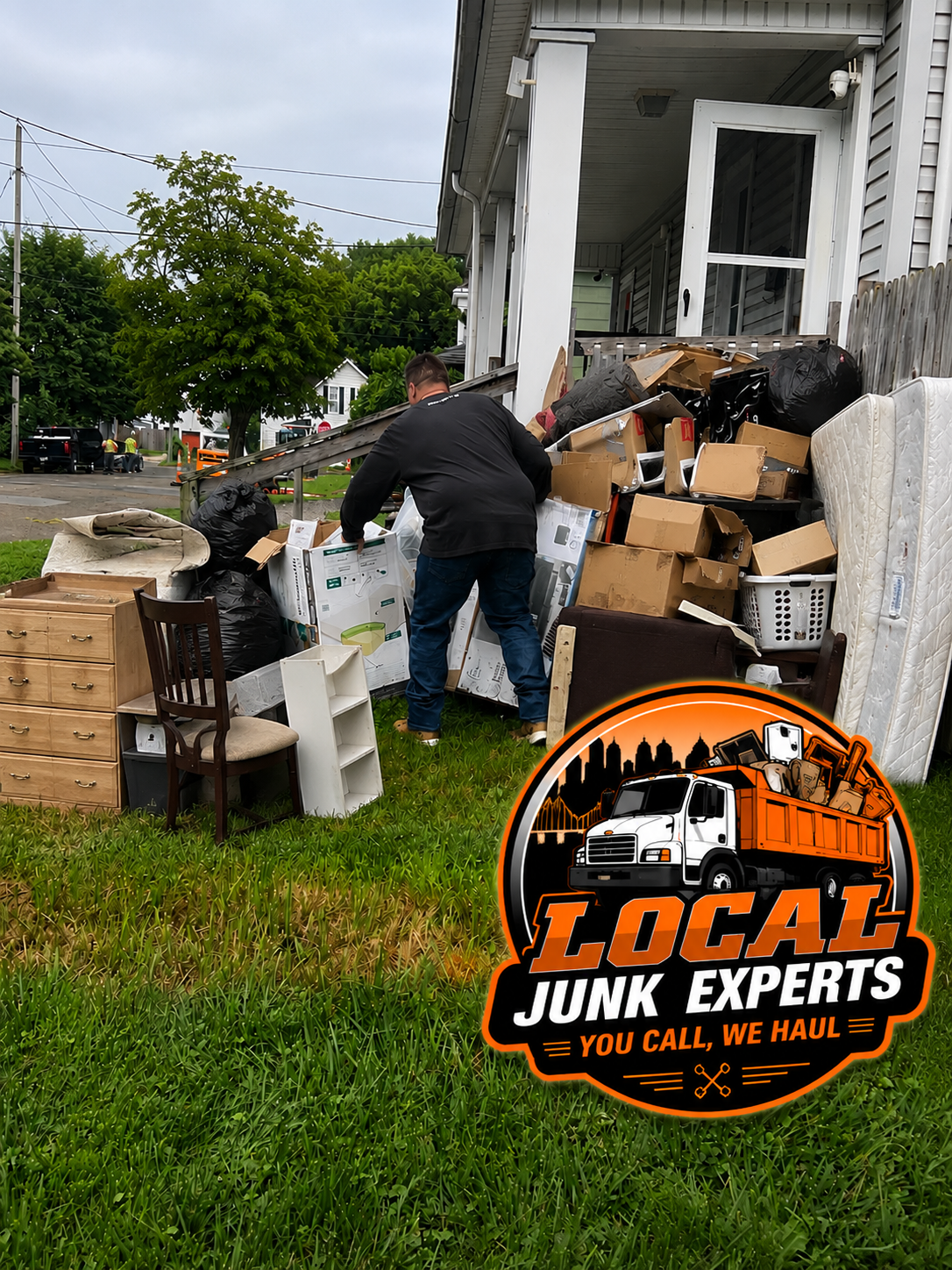 A man disposing of a large pile of household junk, including furniture and cardboard boxes, outside a house. The scene includes an overcast sky, green grass, and neighboring houses. There is a logo for a junk removal service named 'Local Junk Experts' with the tagline 'You call, we haul' in the bottom right corner.