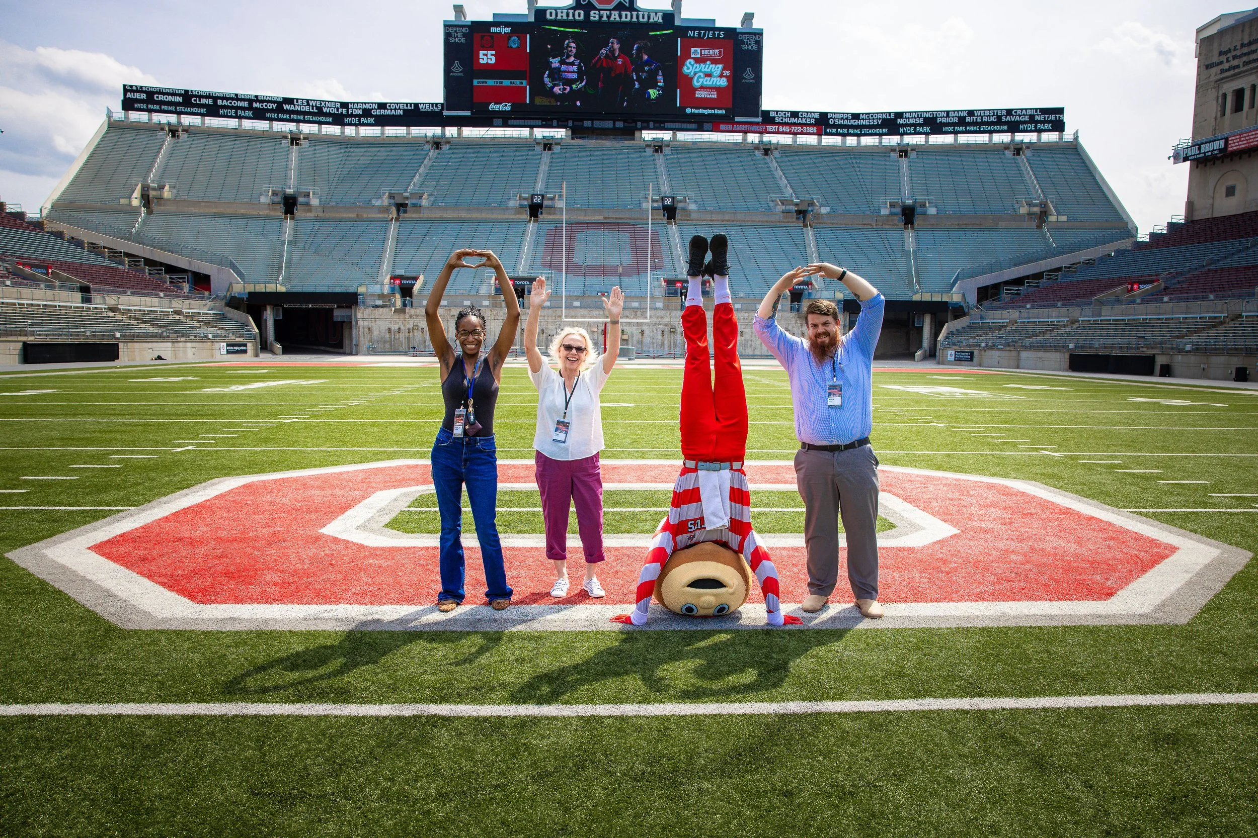 Ohio Stadium football field with empty stands and scoreboard in the background. The mascot is lying on the ground in front of the group, upside down with his head near the center of the field and legs toward the sky. The people are smiling, with two women on the left and two men on the right, one of whom is making a heart shape with his hands. The mascot is wearing a red and white striped jersey.