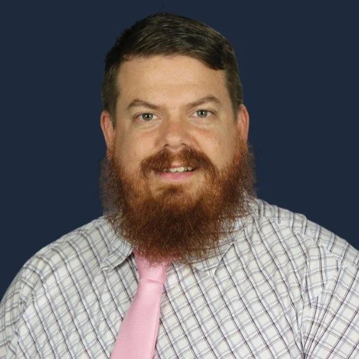Portrait of a bearded man with short brown hair, wearing a checkered shirt and a light pink tie, against a dark blue background.