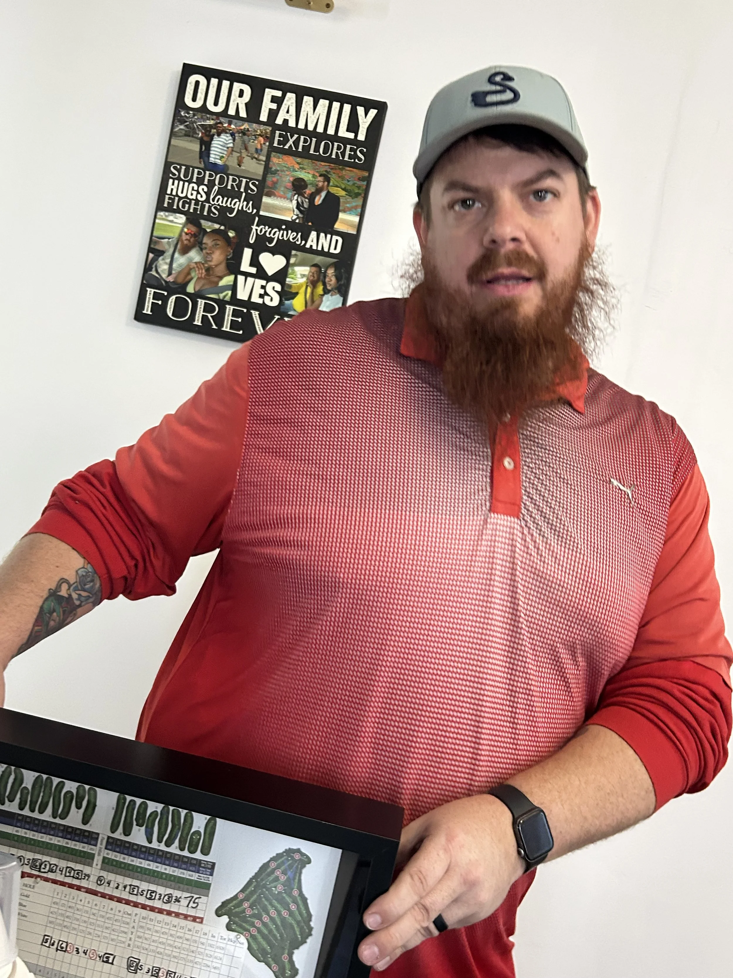 A man with a beard and tattoos on his left forearm wearing a gray baseball cap and a red shirt, holding a golf score sheet or guide with a mini-golf map in front of him, standing in front of a white wall with a black frame reading 'Our Family Explores' with photos and messages about family support, laughter, love, and forgiveness.