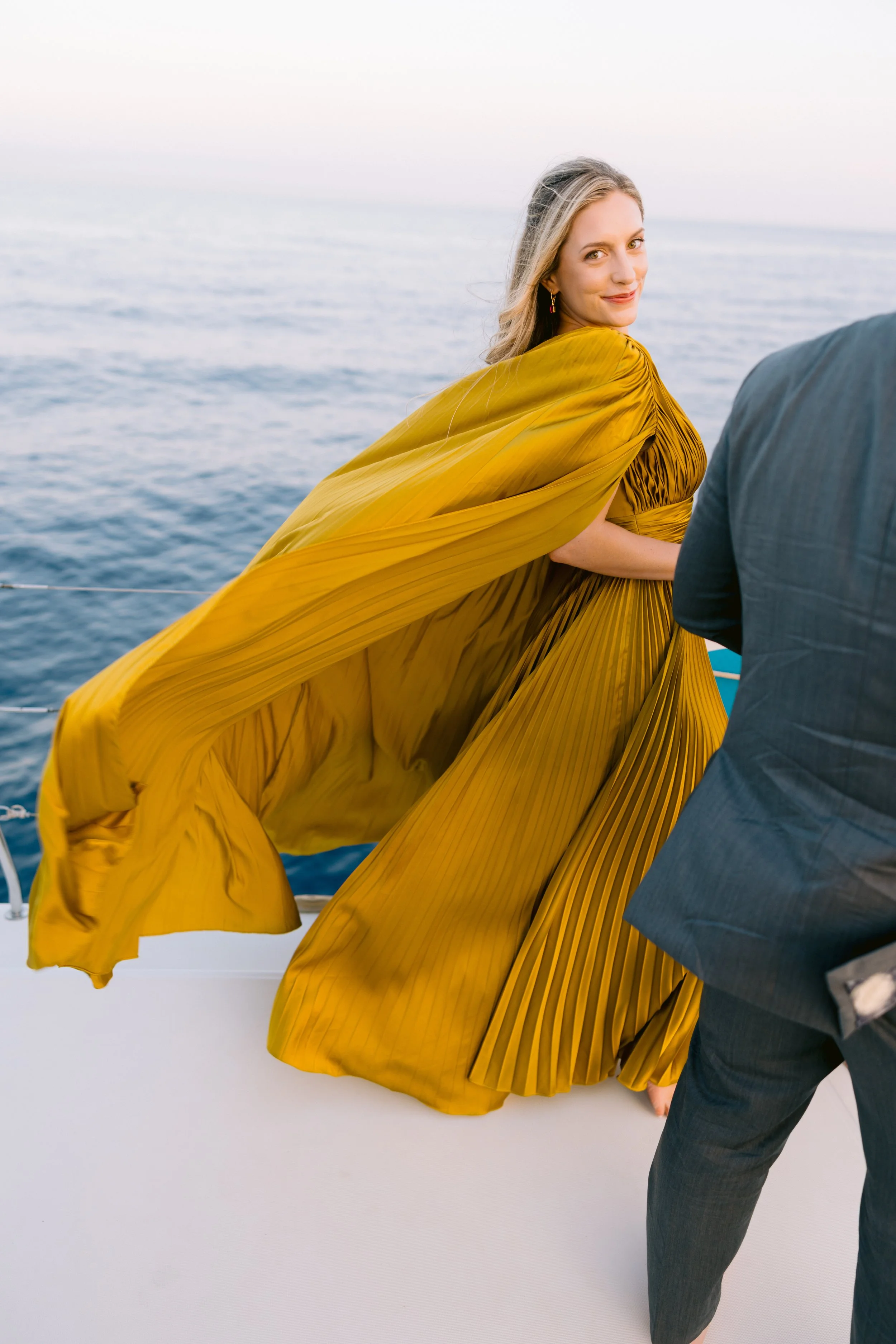 A woman in a yellow pleated dress on a boat, smiling and looking over her shoulder, with the sea in the background.