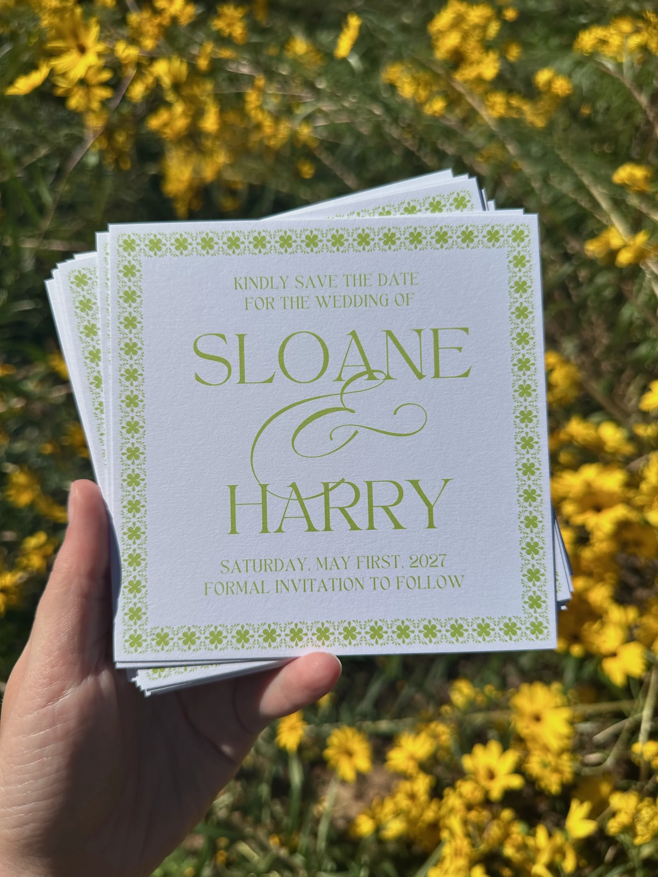 A person is holding a wedding save-the-date card for Sloane and Harry, scheduled for Saturday, May 1st, 2027, outdoors in a field of yellow flowers.