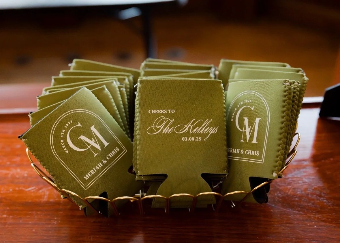 Green napkins with wedding details, including initials 'G' and 'M,' date March 8th, 2025, and names Meriah and Chris, in a gold wire basket on a wooden table.