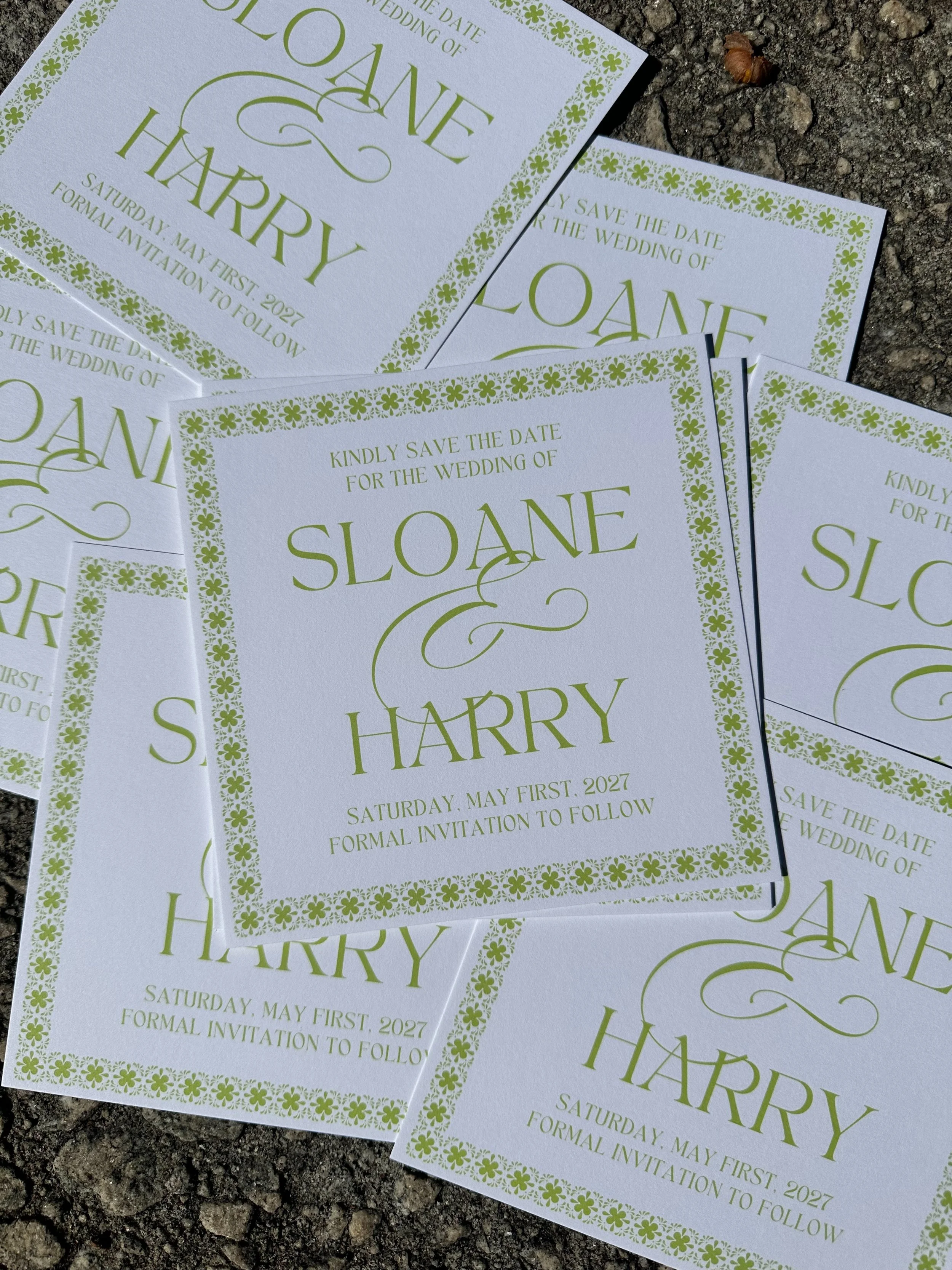 Multiple white wedding save the date cards with green floral border and text, laying on rough ground. Momentos Vivere.