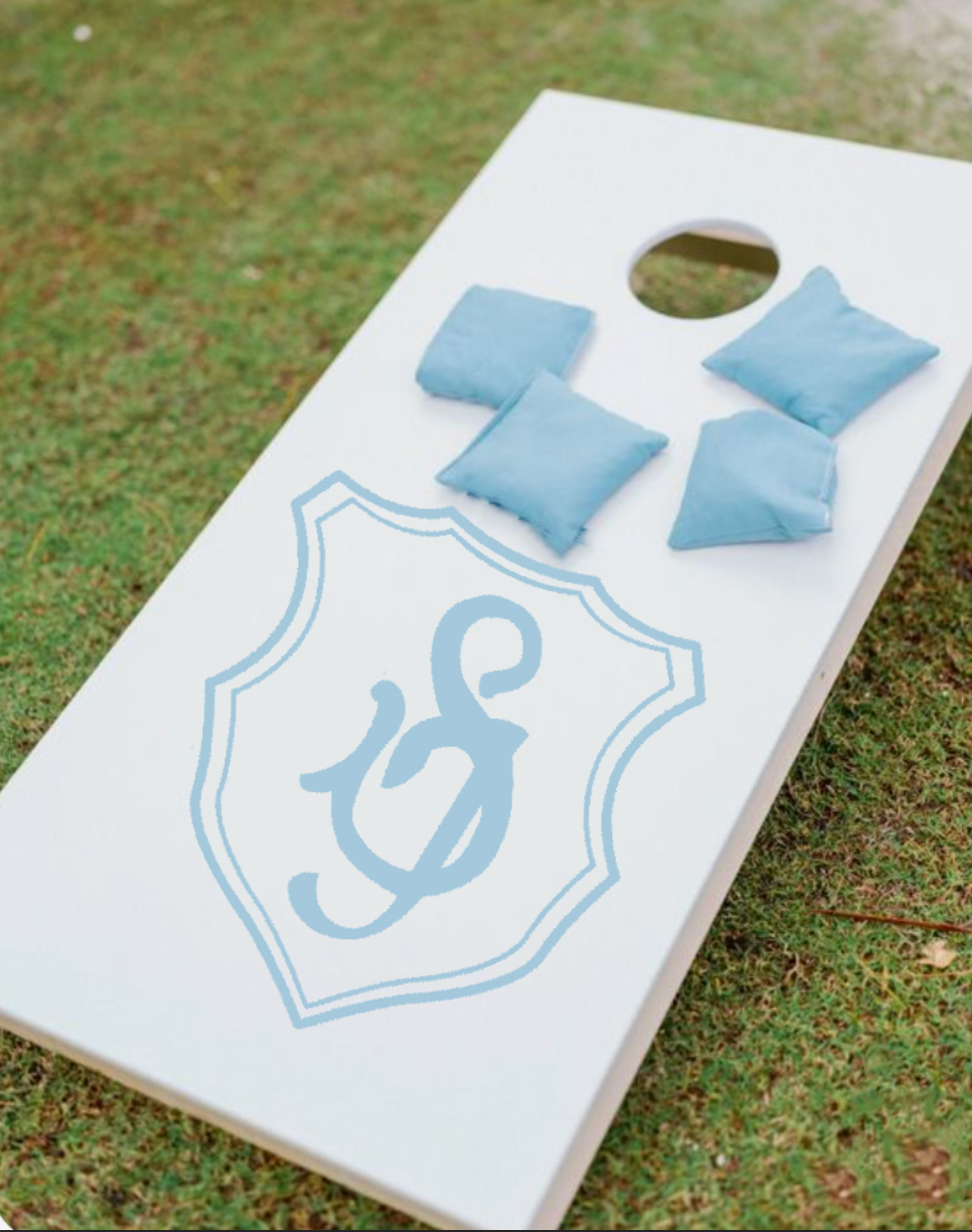 A cornhole game board with blue bean bags on it, set outdoors on grass. The board has a light custom crest bag throw board coastal wedding feel.