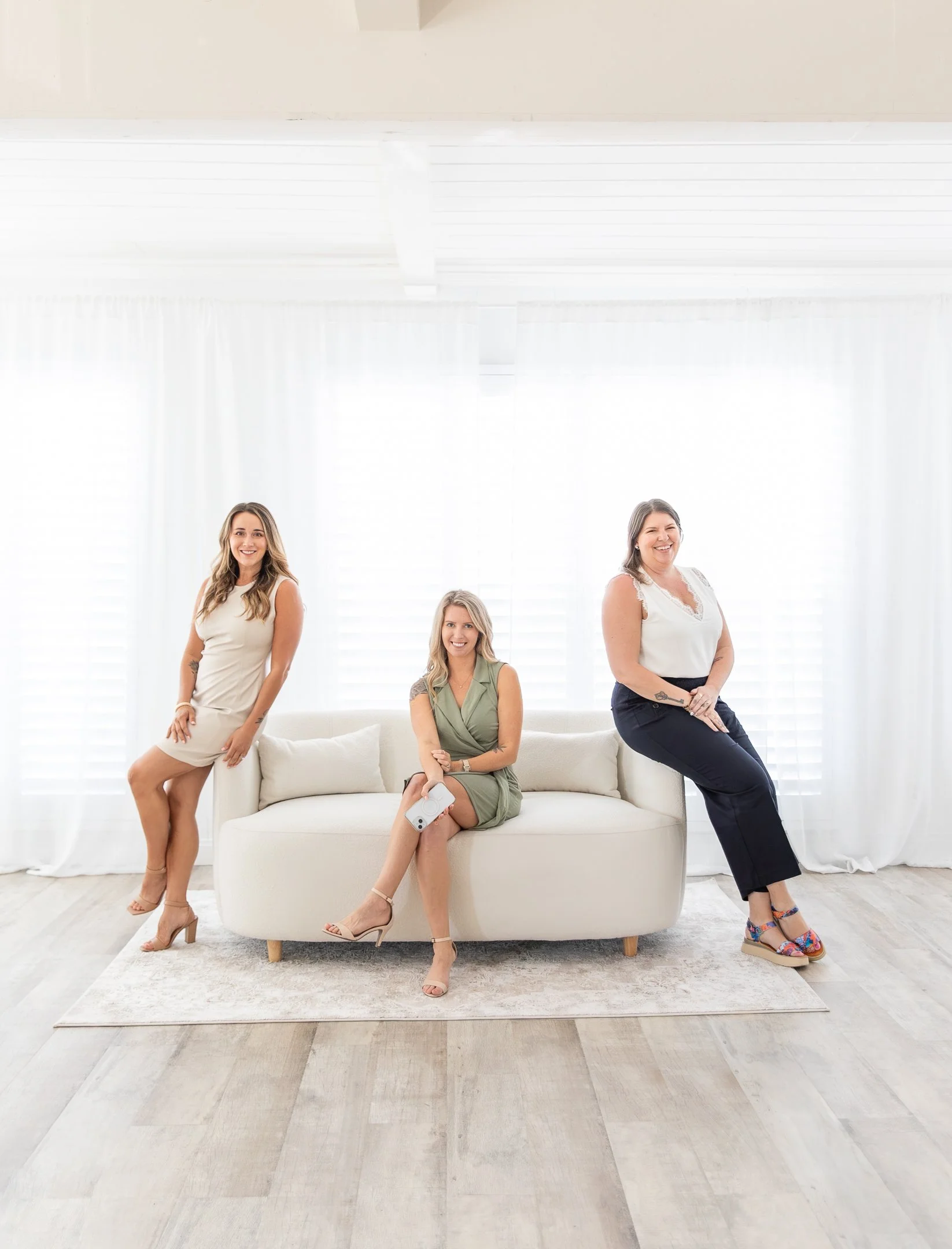 Three women sitting and standing on a white sofa in a bright, white, minimalist room with white curtains and hardwood floor.
