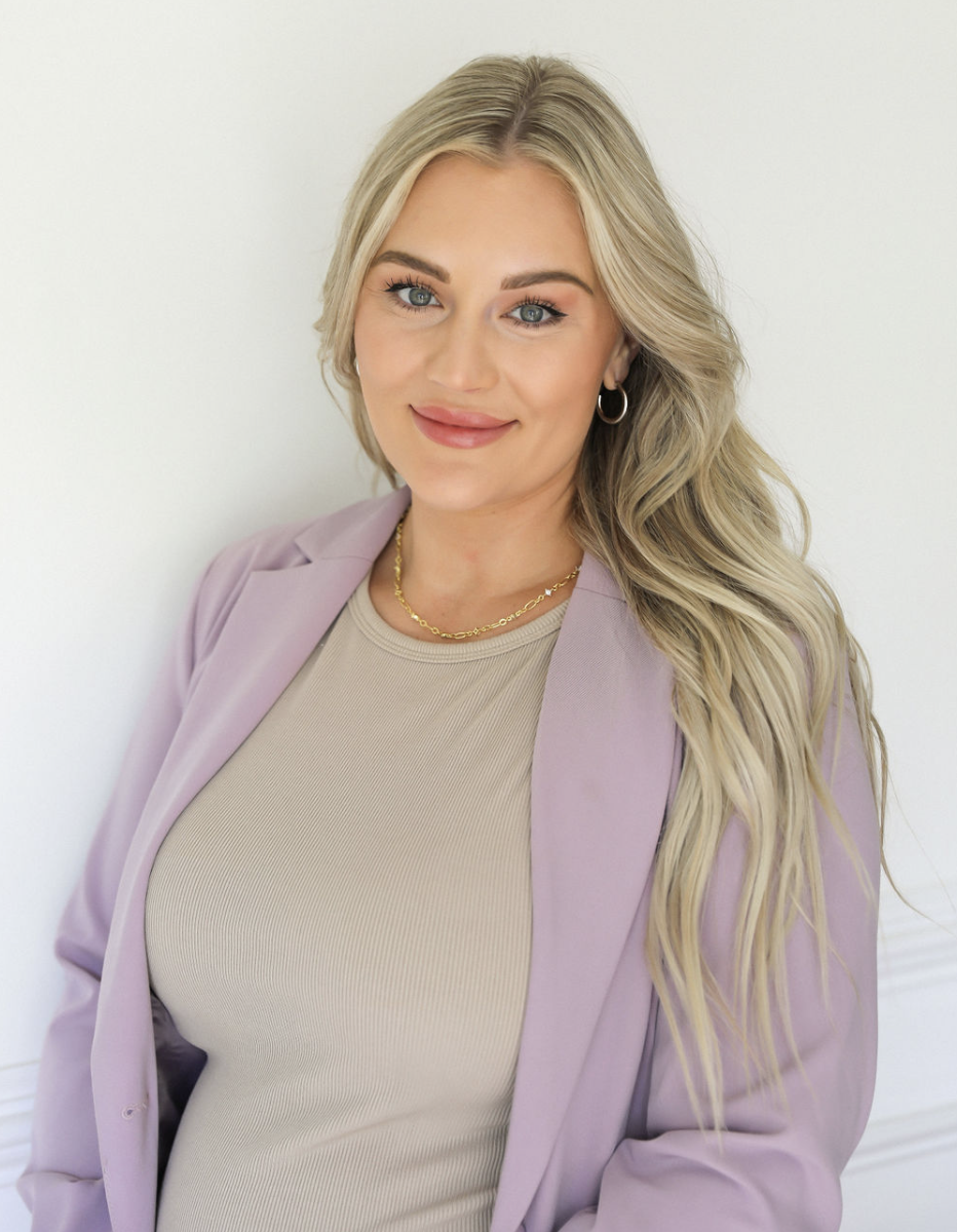Portrait of a young woman with blonde hair, blue eyes, wearing a beige top, a light purple blazer, gold jewelry, and hoop earrings, standing against a white wall.