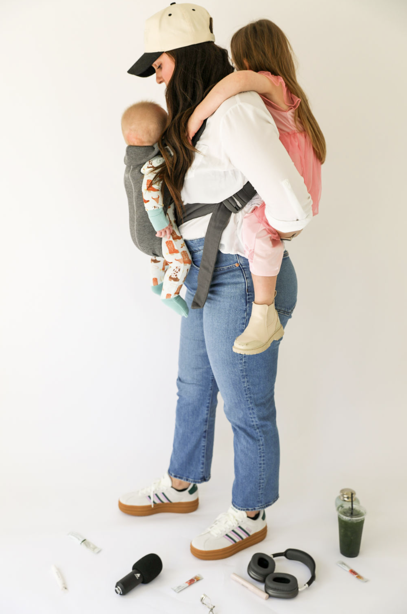 A woman carrying a young girl on her back and a baby in a front carrier, standing on a white floor with various items scattered around, including a green drink, headphones, a small flashlight, and candies.