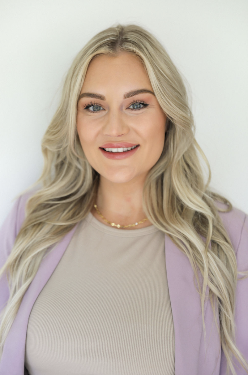 A young woman with long, wavy blonde hair and blue eyes smiling at the camera, wearing a beige top, a light purple blazer, and a gold necklace, against a plain white background.