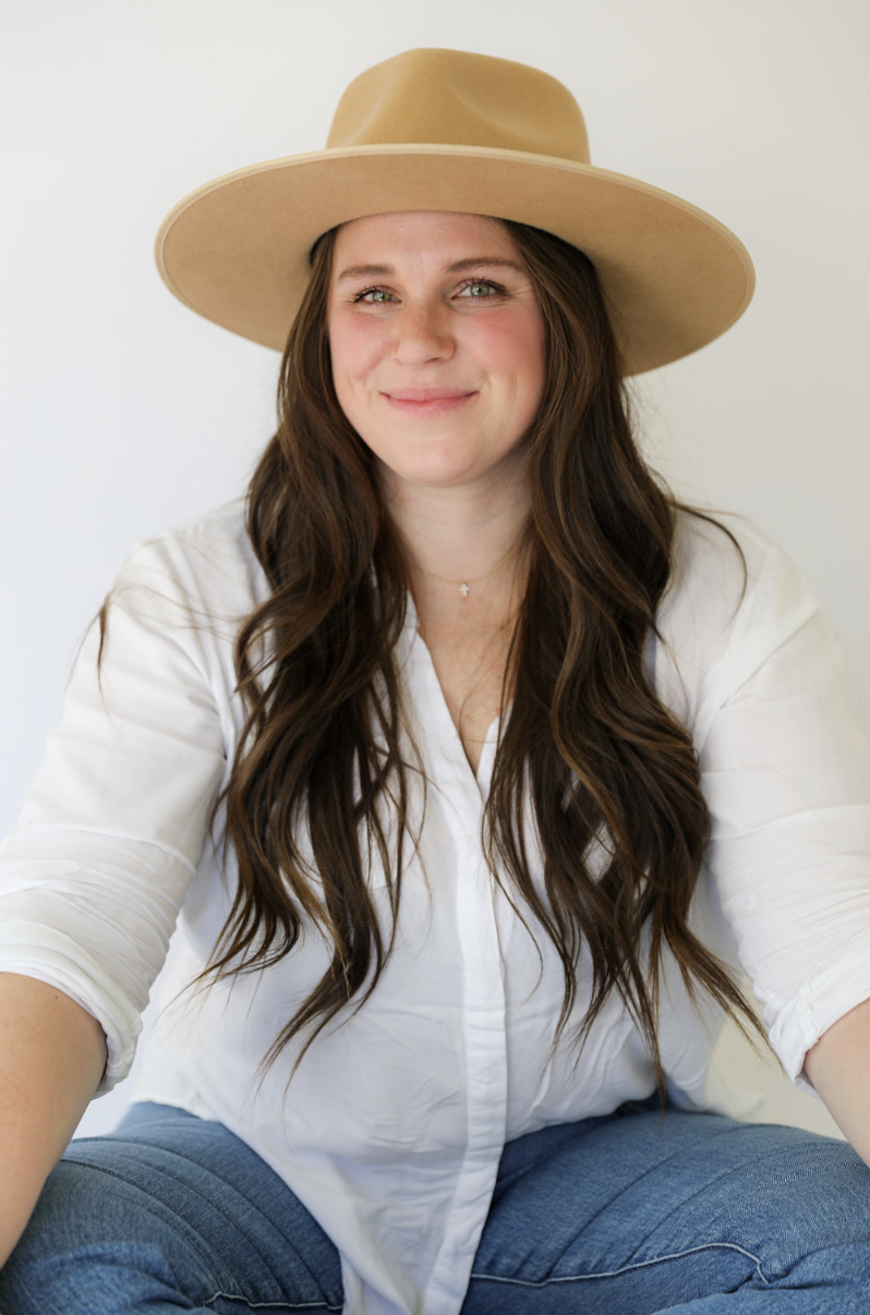 A woman with long brown hair wearing a beige wide-brimmed hat, a white button-up shirt, and blue jeans, sitting against a plain light background.