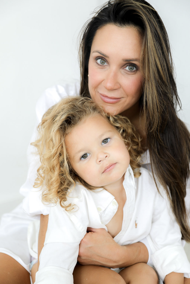 A woman with long dark hair and striking blue eyes holding a young girl with curly blonde hair and blue eyes, both dressed in white, in a bright, neutral setting.