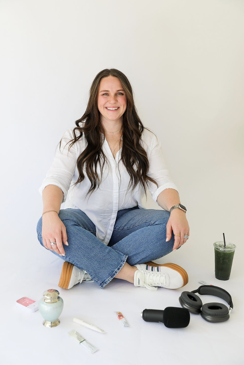A woman with long brown hair, wearing a white shirt and blue jeans, sitting cross-legged on a white surface, smiling, with accessories and drinks around her.