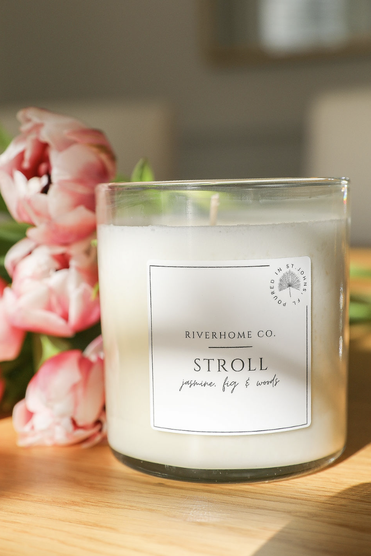 A white candle in a glass jar labeled 'Riverhome Co. Stroll jasmine, fig & woods' sits on a wooden surface next to pink tulips with green leaves.