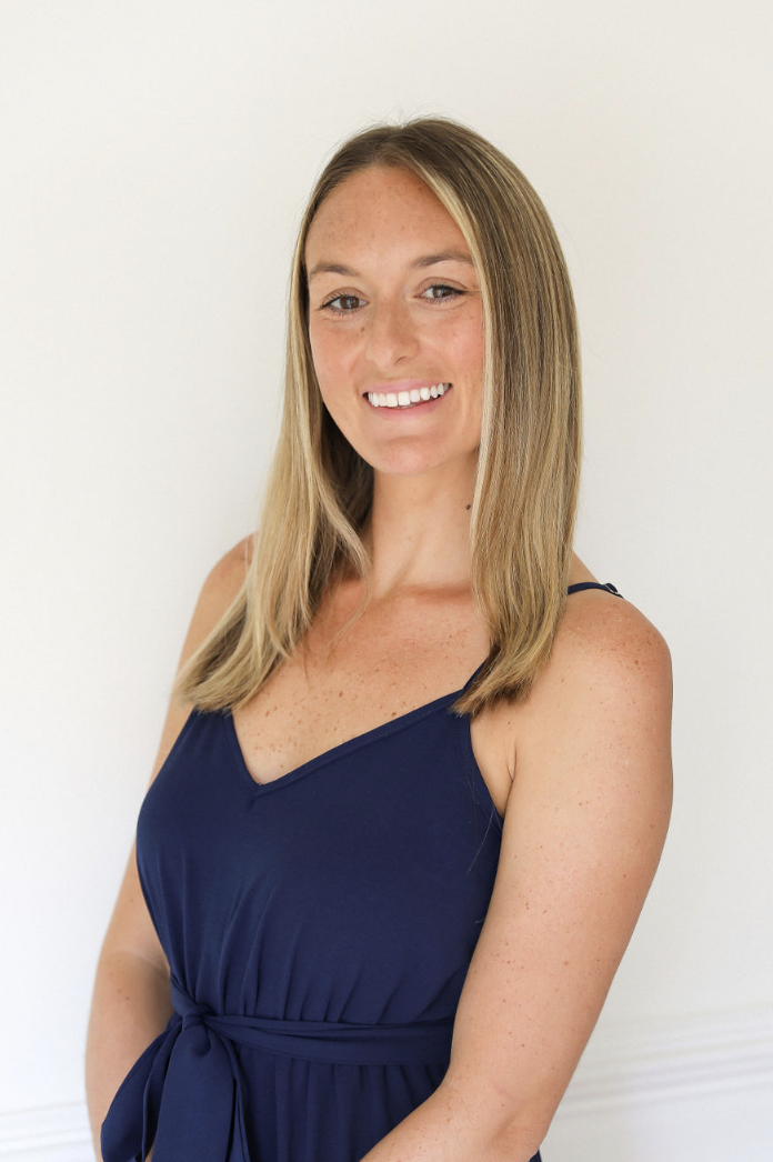 A woman with long blonde hair wearing a navy blue sleeveless dress smiling at the camera.