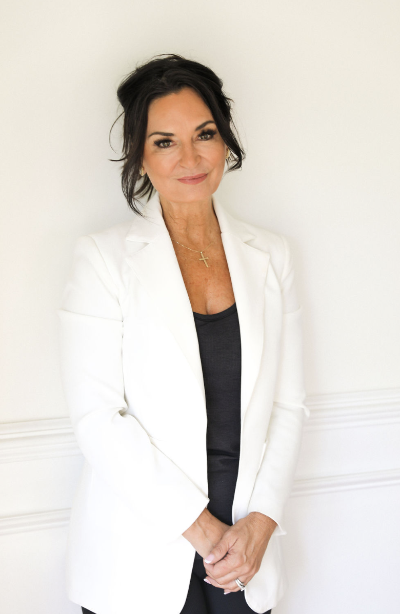 A woman with short dark hair wearing a white blazer, black top, and a gold cross necklace stands against a white wall.