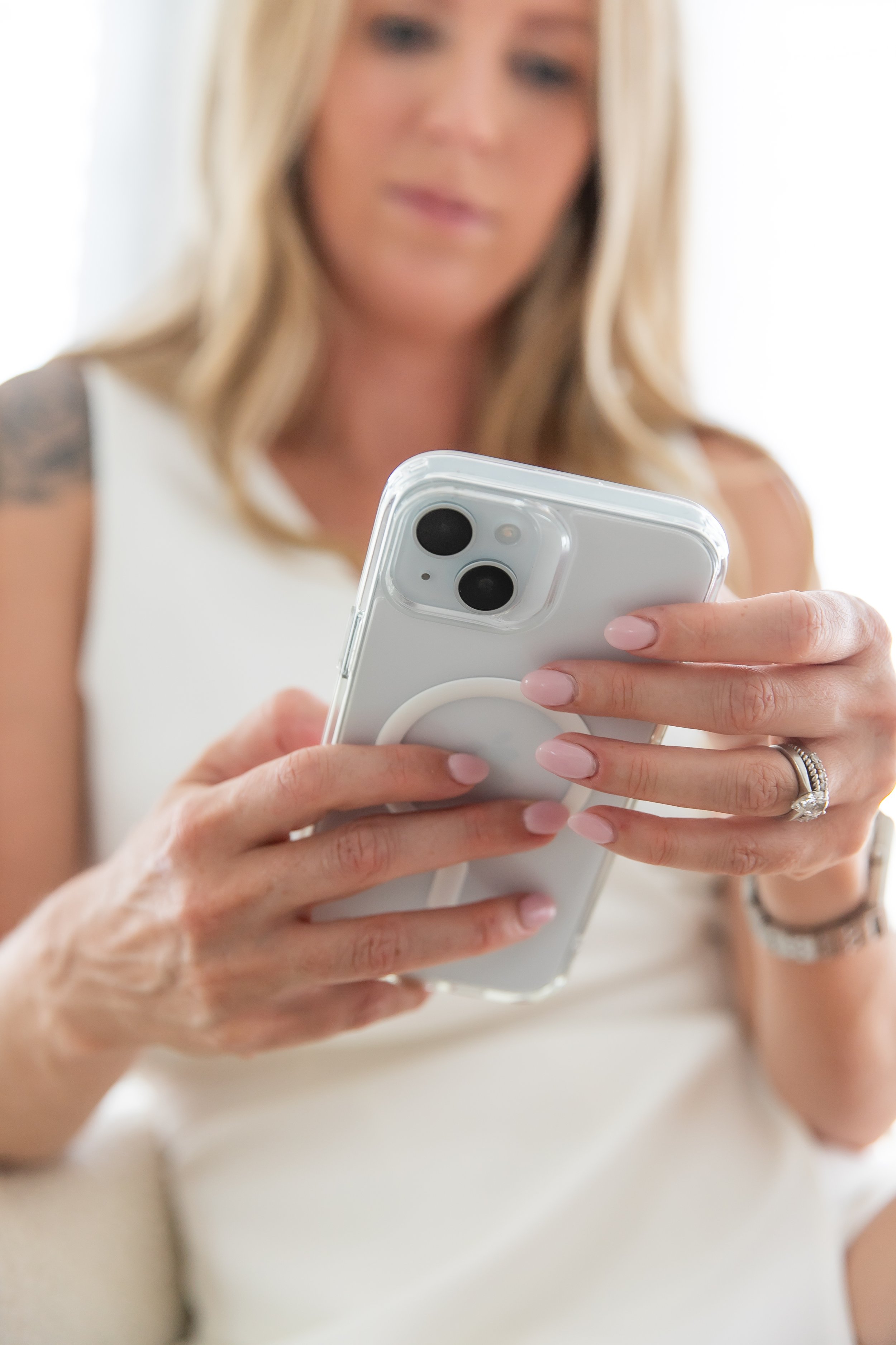 A woman with blonde hair, wearing a white sleeveless top, holding a smartphone with a clear case and pink nail polish, appears to be using the phone.
