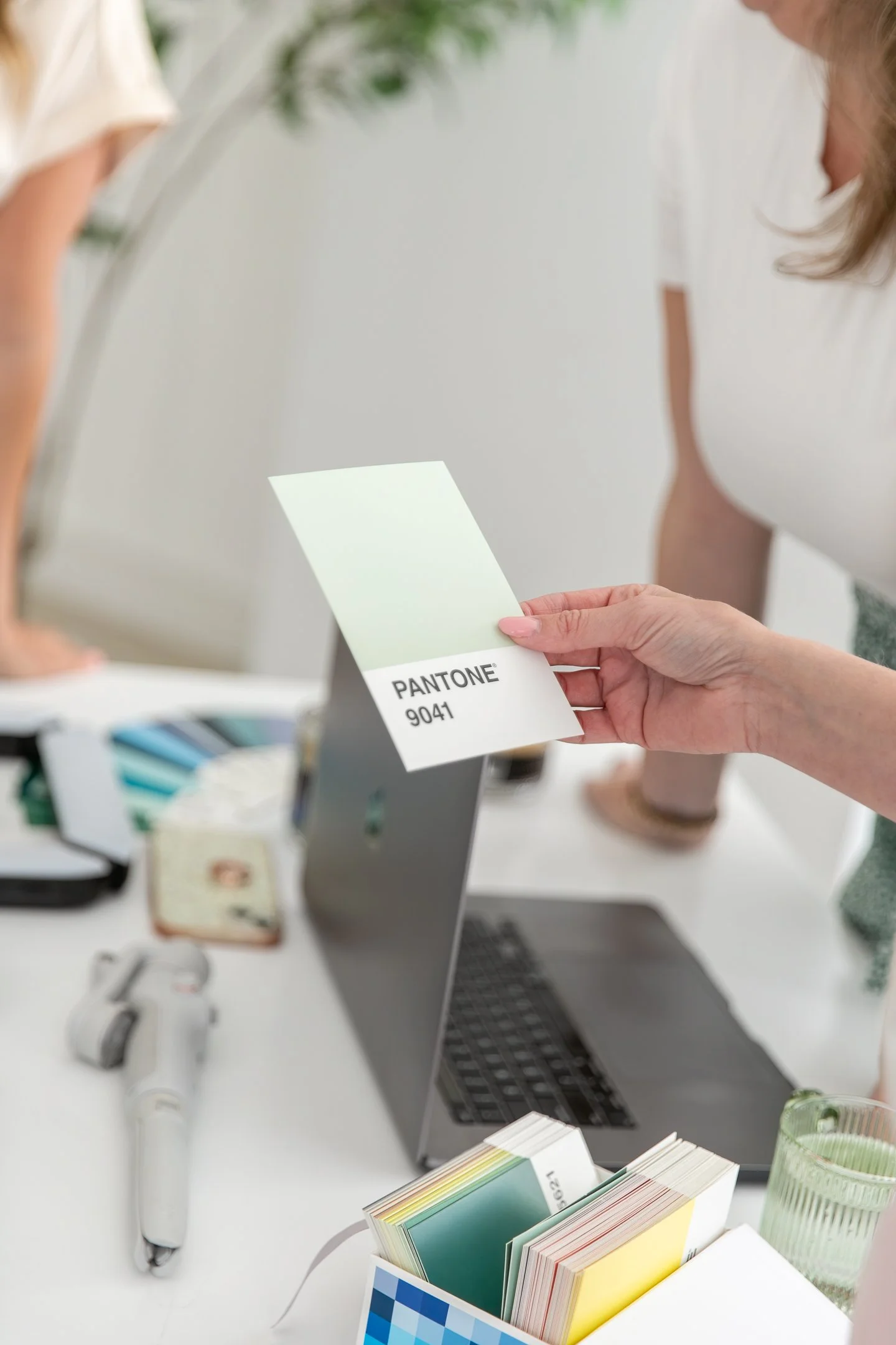 Person holding a Pantone color sample card labeled 9041 in front of a mirror, on a desk with color swatches, a laptop, a glass of green drink, and other items.