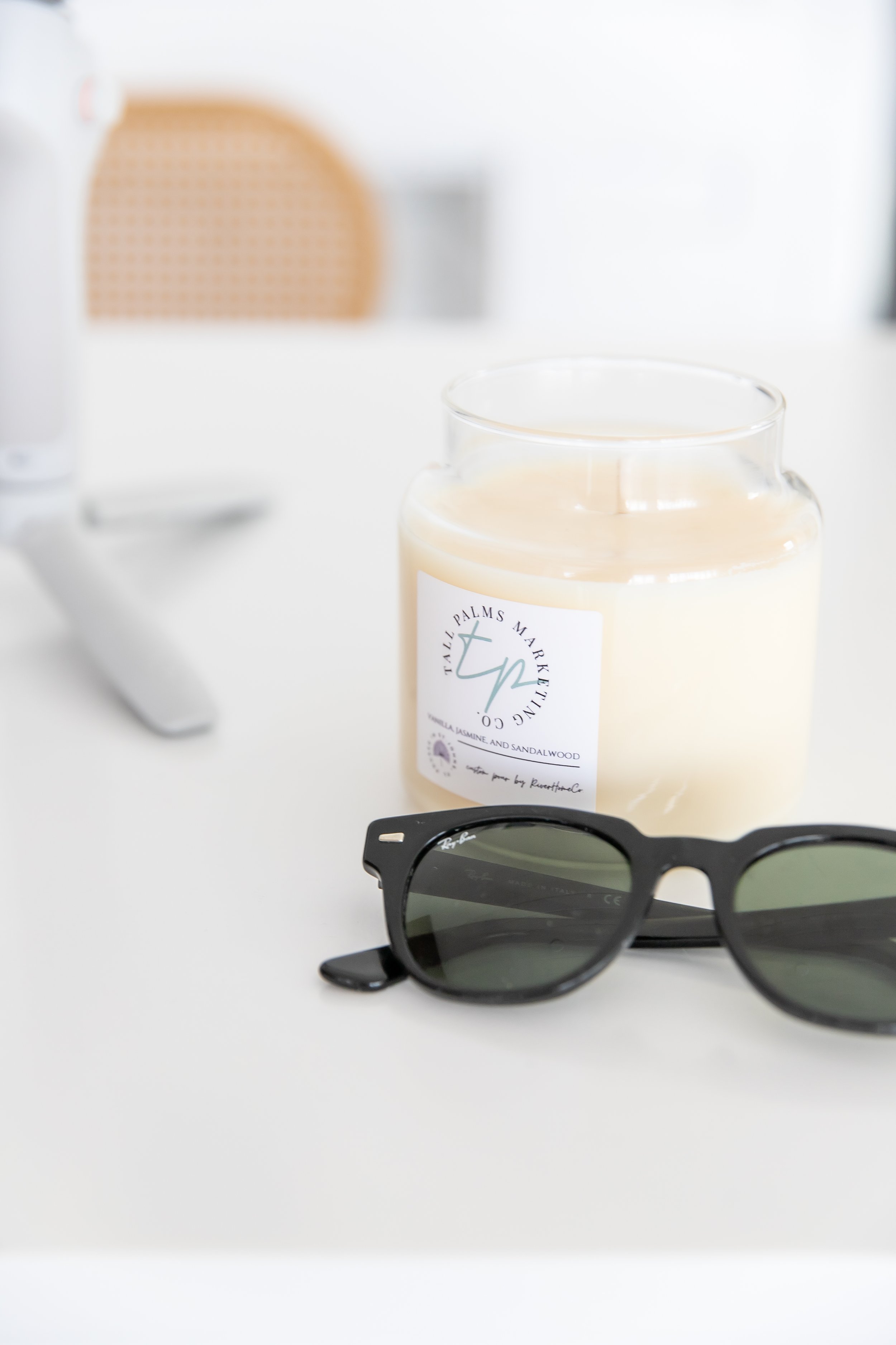 A pair of black sunglasses, a white candle with a label, and a blurred background on a white surface.