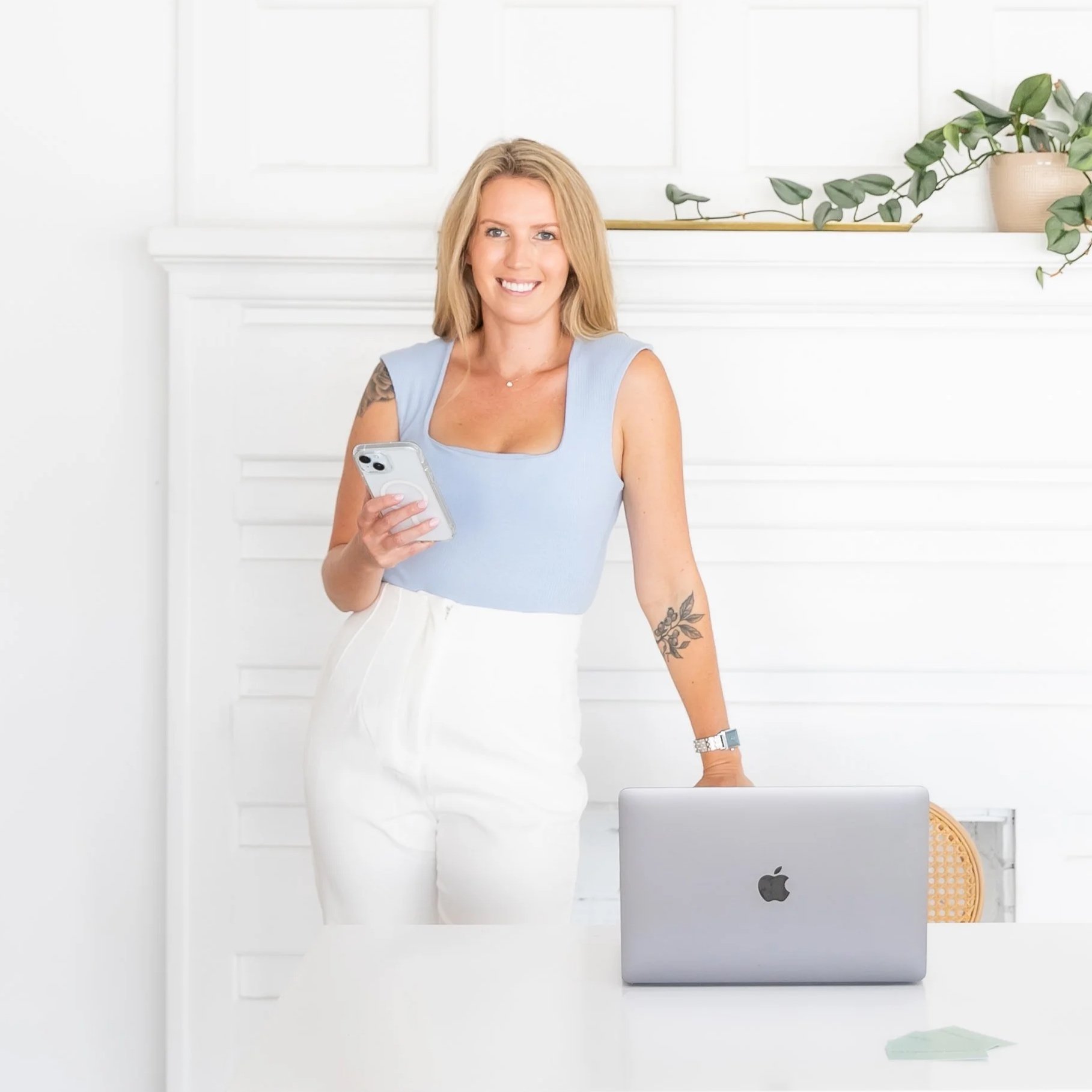 A woman with blonde hair, wearing a light blue sleeveless top and white pants, standing in a bright, white room, holding a smartphone, with a silver laptop on a white table in front of her, and a potted plant on a white shelf behind her.