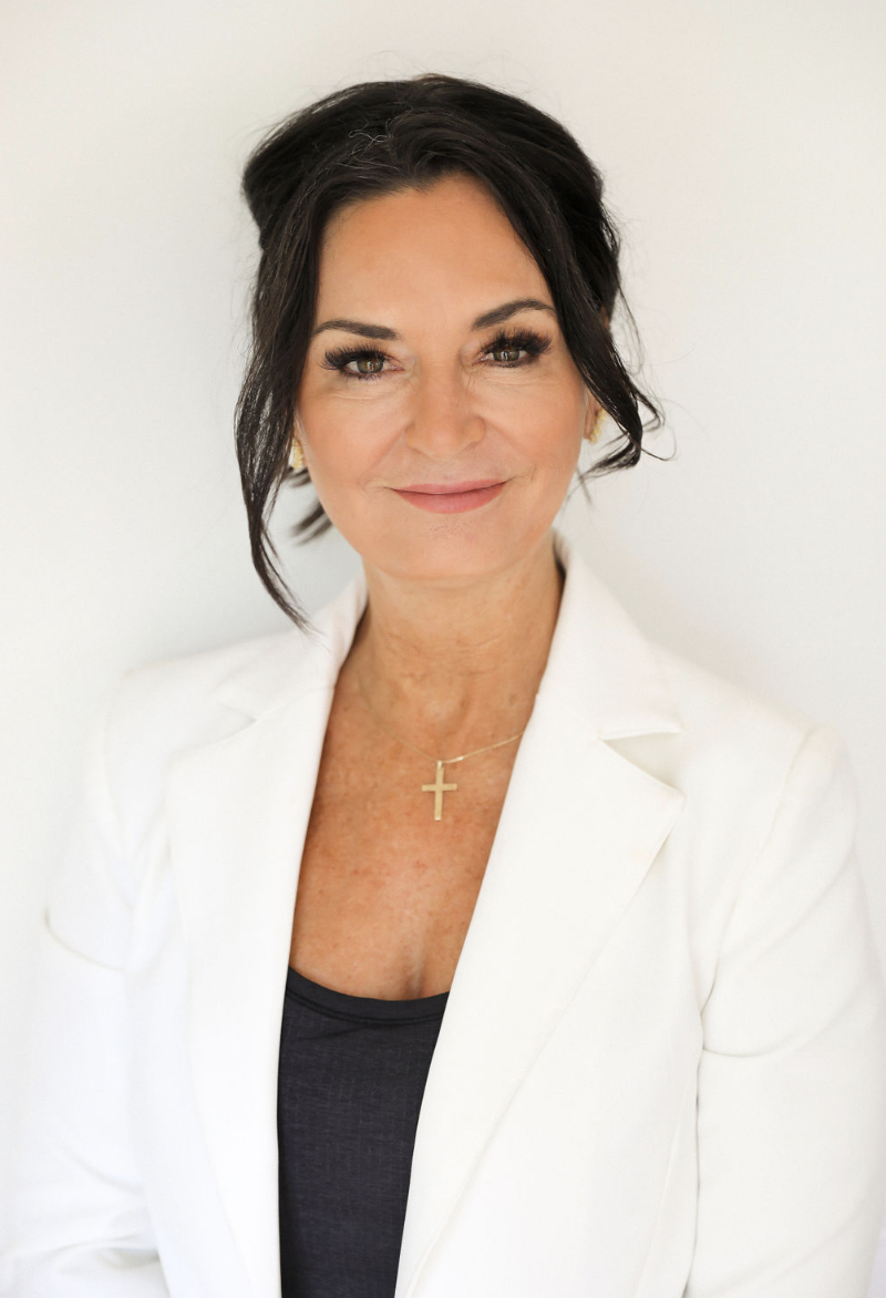 A woman with short dark hair, wearing a white blazer, black top, and a gold cross necklace, smiling softly against a plain white background.