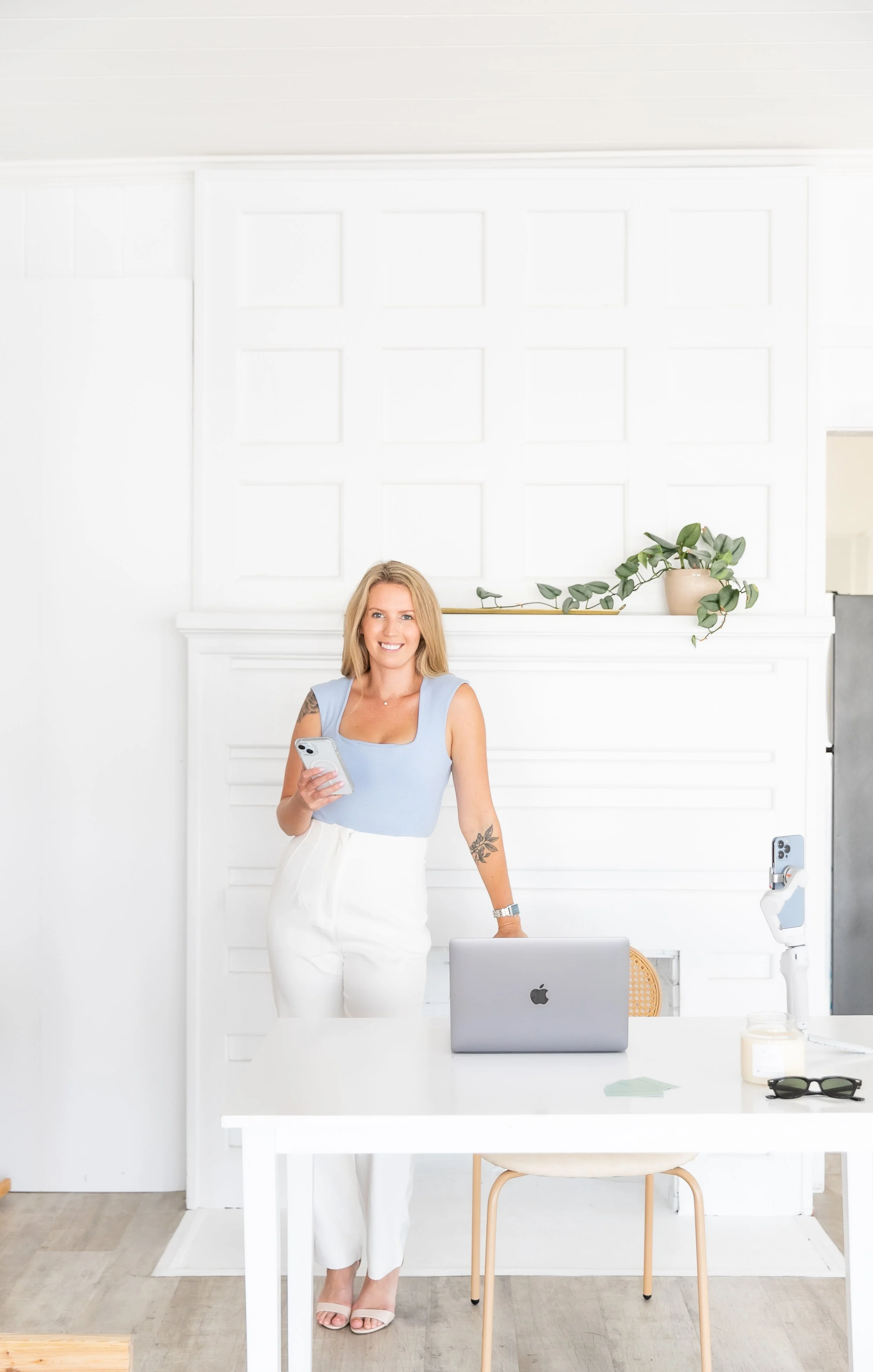 A woman standing behind a white table with a laptop, holding a smartphone, smiling in a bright, minimalist room with white walls and a plant.