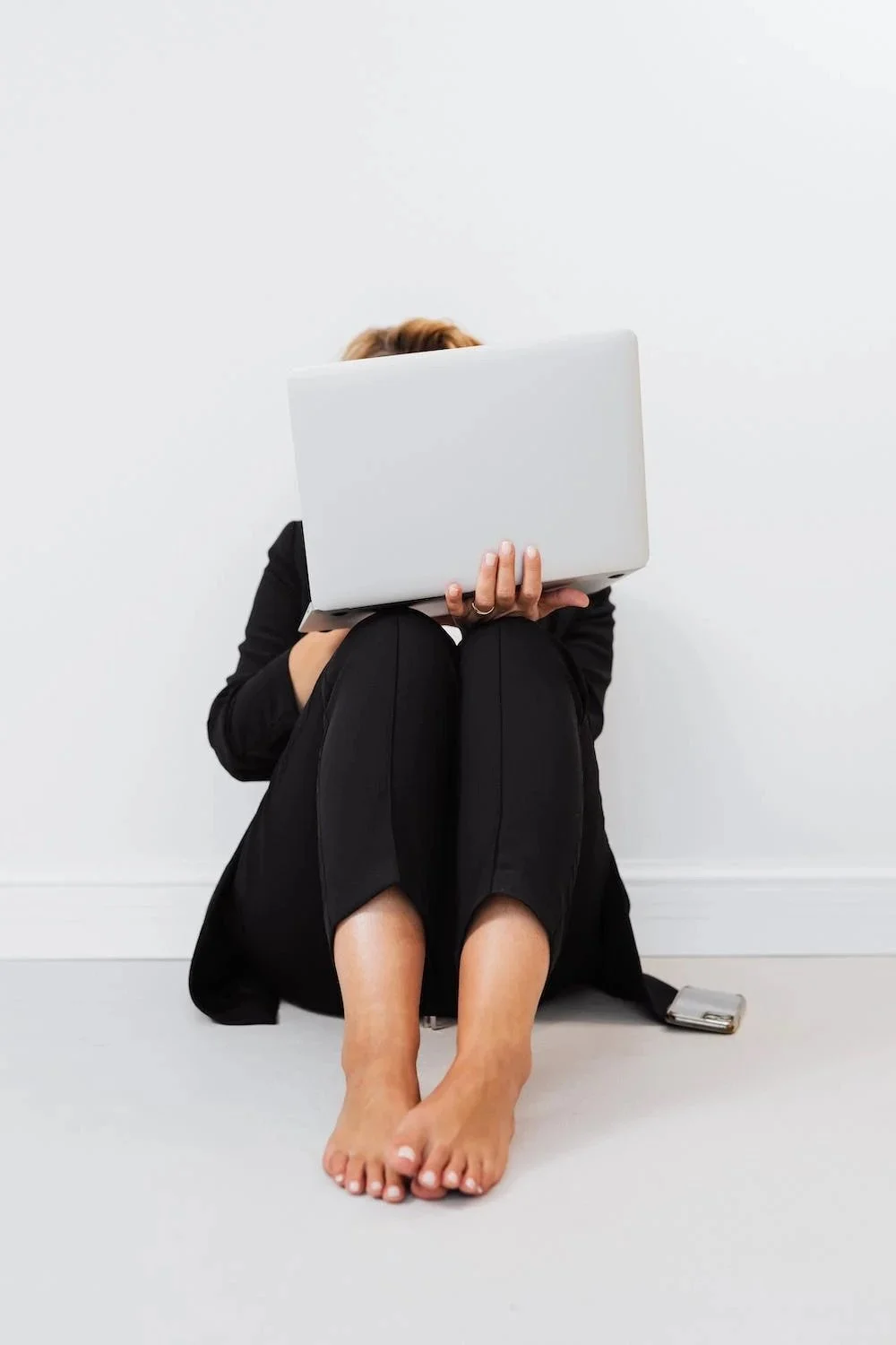 Person sitting on the floor with legs crossed, holding a laptop in front of their face, wearing black pants and a dark blazer, with a smartphone on the floor beside them, against a plain white wall.