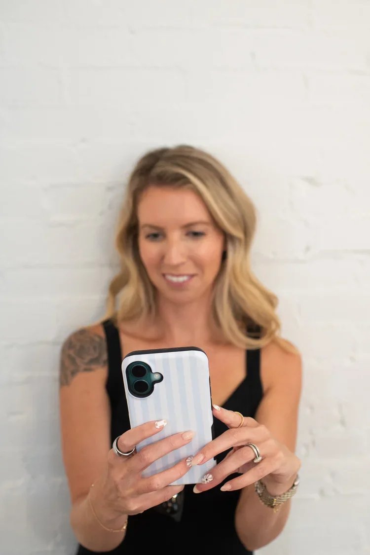 Woman with blonde hair taking a selfie with her smartphone, standing against a white brick wall.