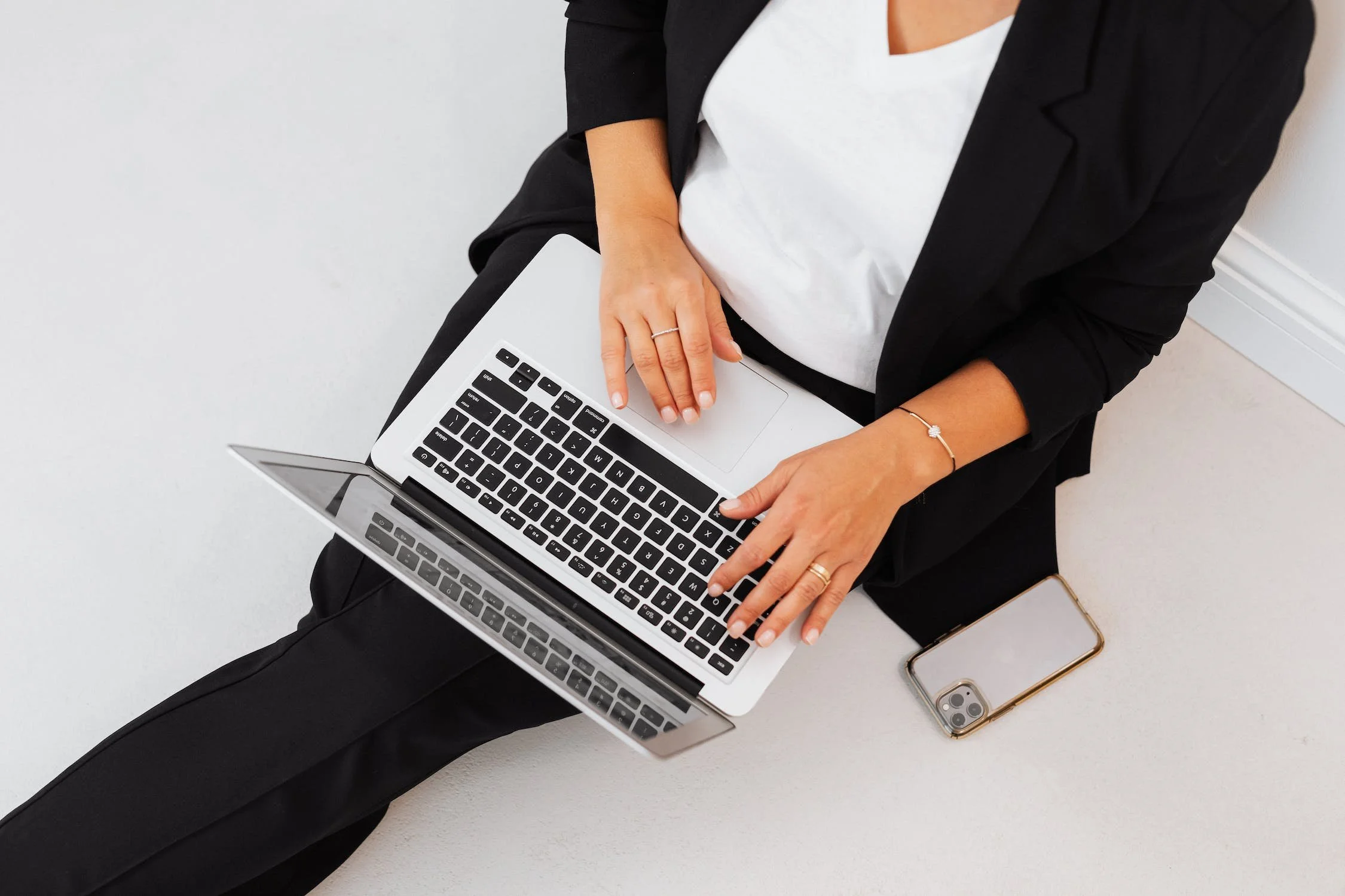 A woman sitting on the floor using a silver laptop with a black keyboard, wearing a black blazer and black pants, with a smartphone placed next to her on the floor.