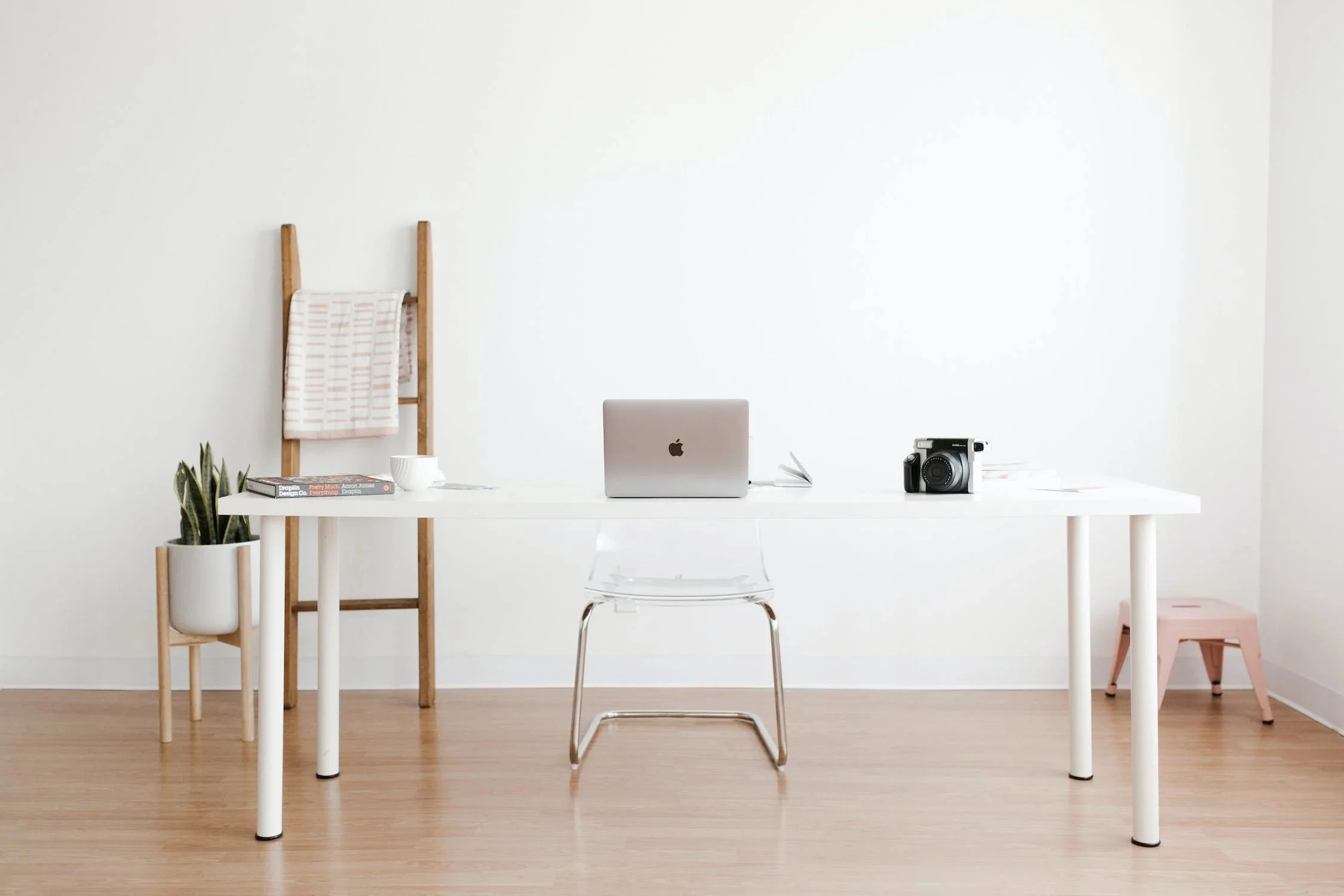 Minimalist white desk in a bright room with a laptop, a camera, a stack of books, a teapot and mug, and a plant. A wooden ladder with towels draped over it to the left, and a pink stool to the right.