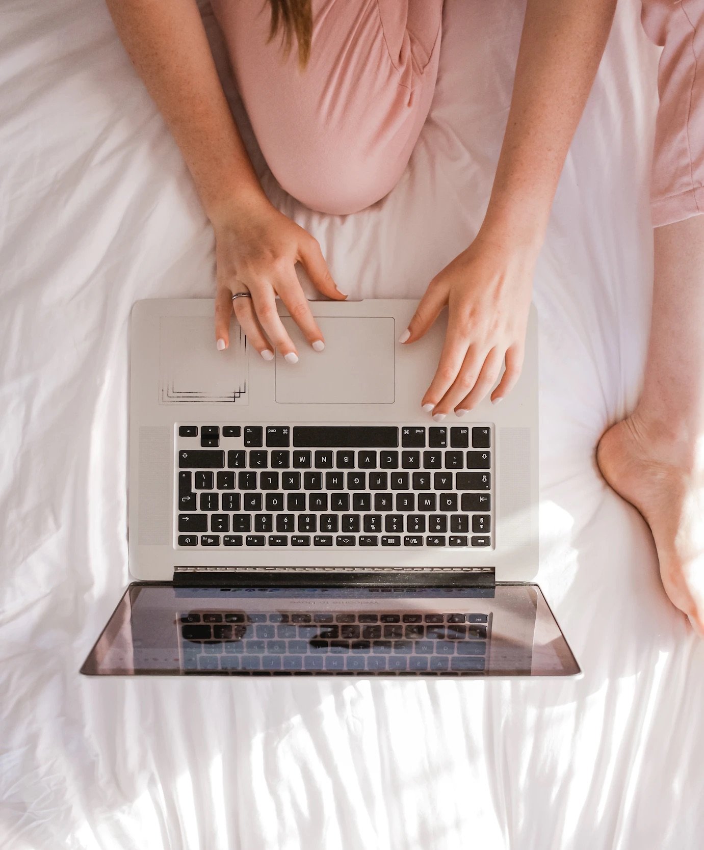 Person sitting on a bed with a silver laptop, wearing pink pajamas, with legs bent and hands on the keyboard.