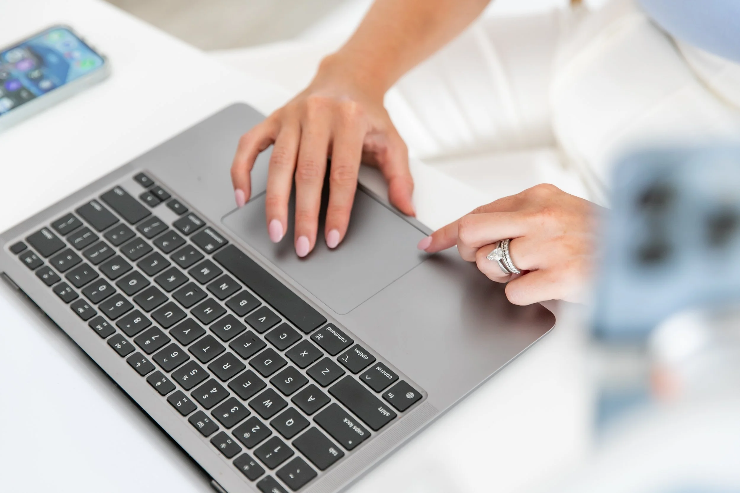 A person's hand with a wedding ring on the ring finger resting on a laptop trackpad, with a smartphone on a white surface nearby.