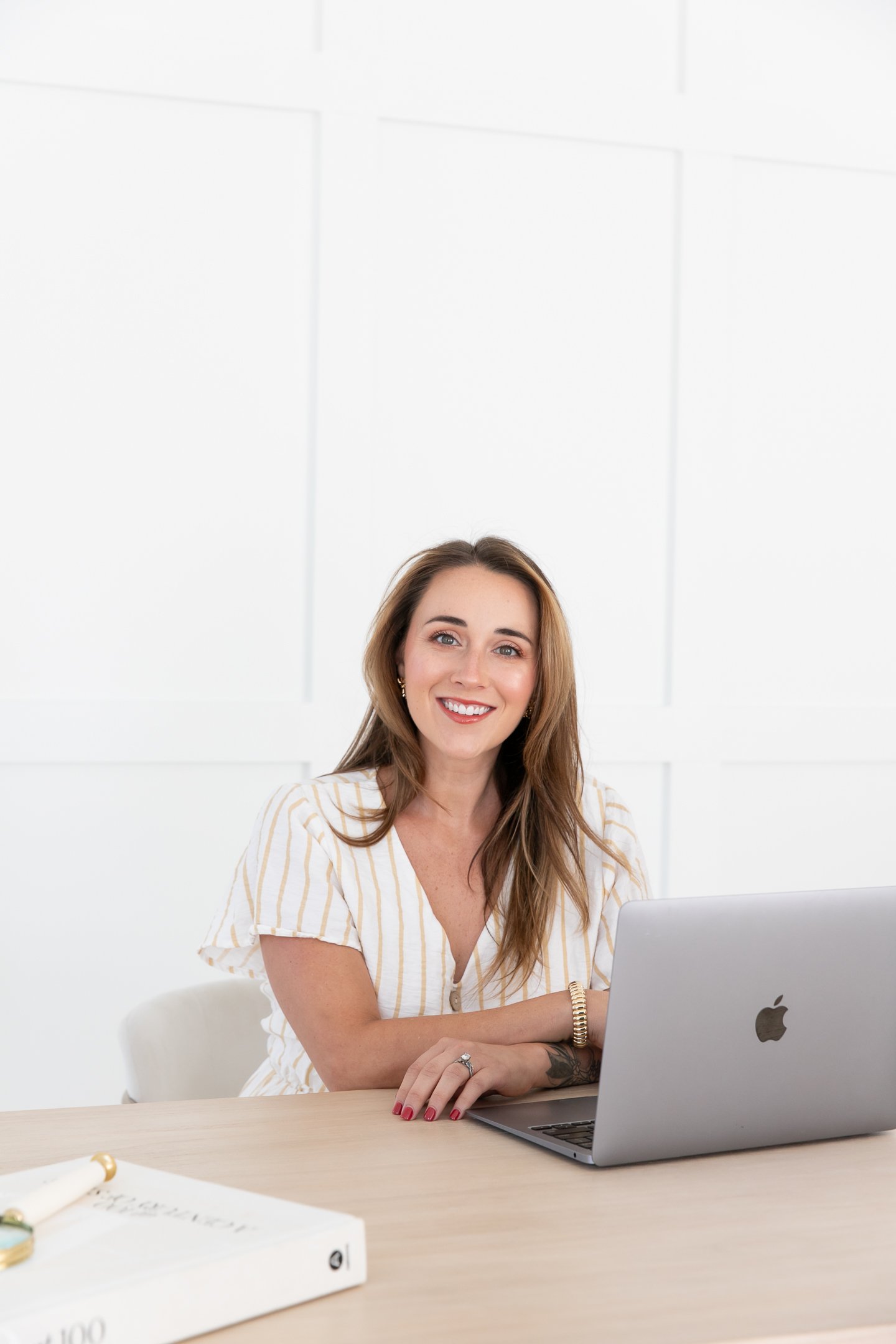 A woman with long brown hair smiling while sitting at a desk with a silver laptop and a white notebook with gold accents.