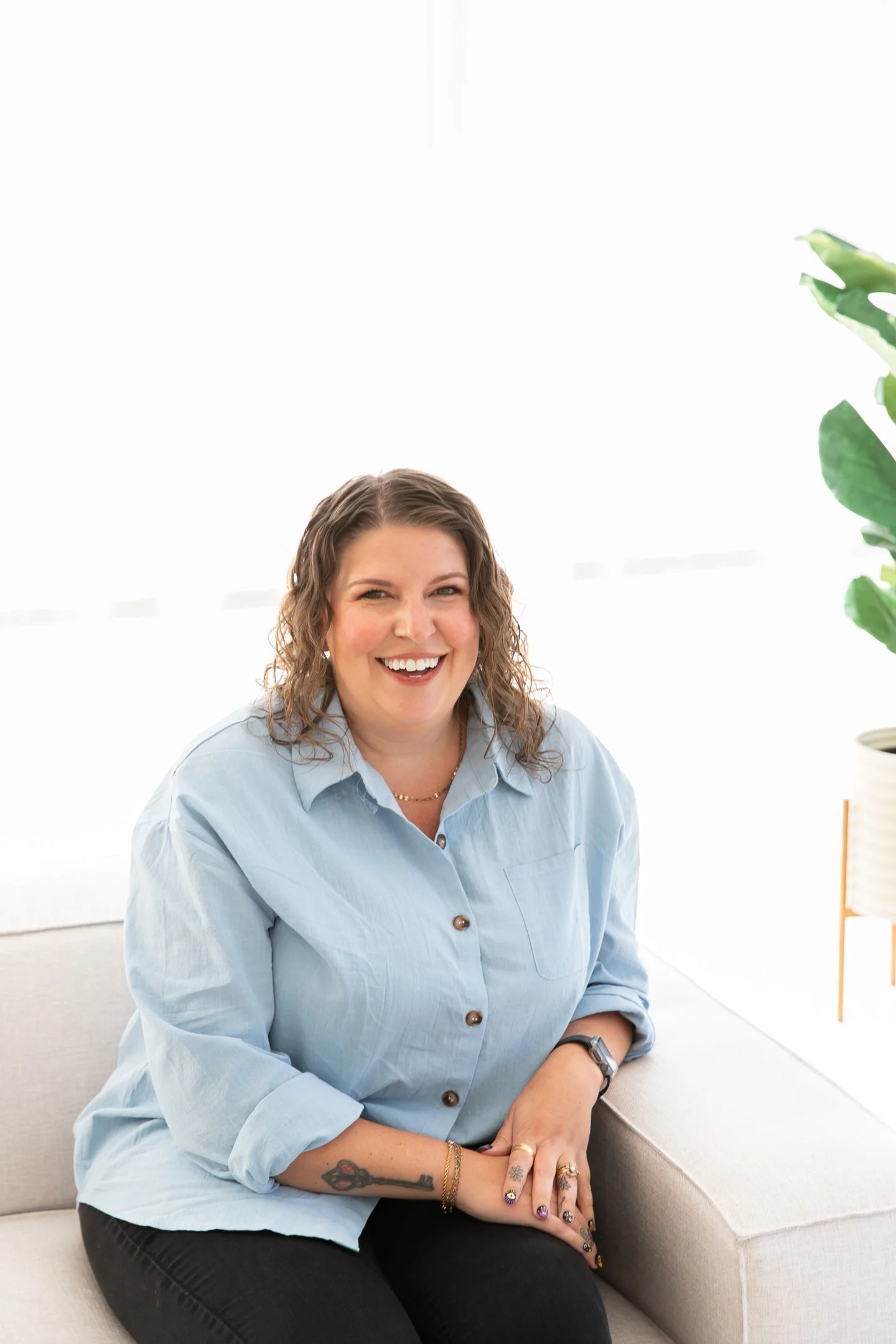 A smiling woman with wavy brown hair sitting on a light-colored sofa in a bright room with white curtains and a plant in the background.