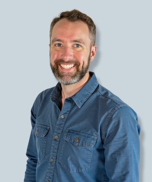 Portrait of a smiling man with short brown hair, beard, wearing a blue denim shirt, standing against a light gray background.