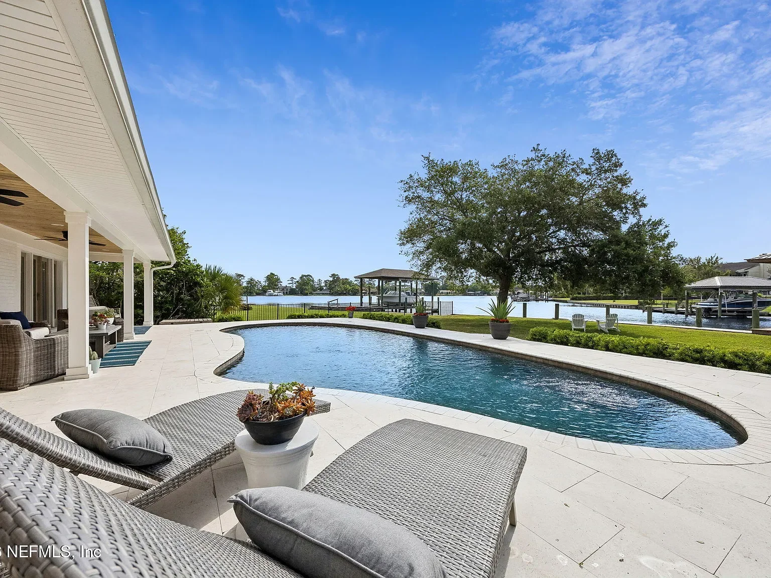 Backyard with a swimming pool overlooking a waterway, with potted plants, lounge chairs, a large tree, and a covered pavilion by the water.