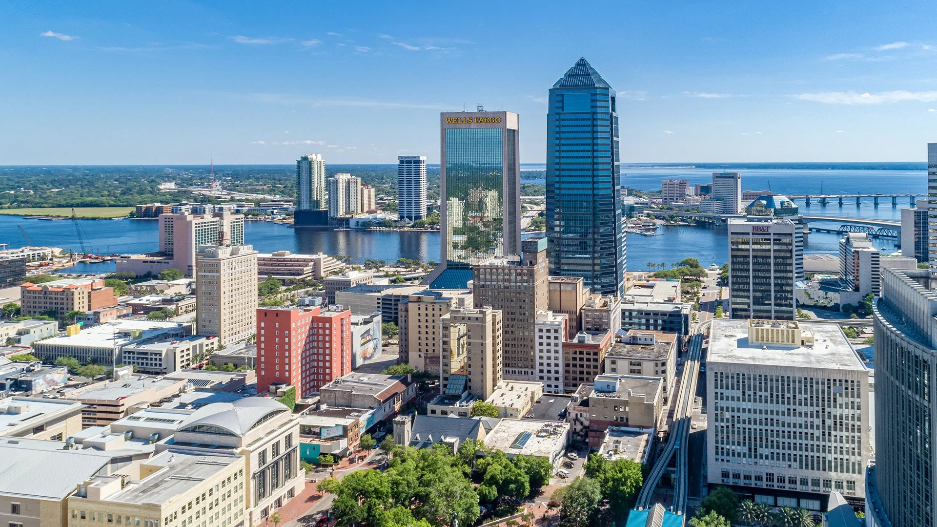 Aerial view of a city skyline with modern high-rise buildings along a river, with bridges crossing the water, under a blue sky with some clouds.