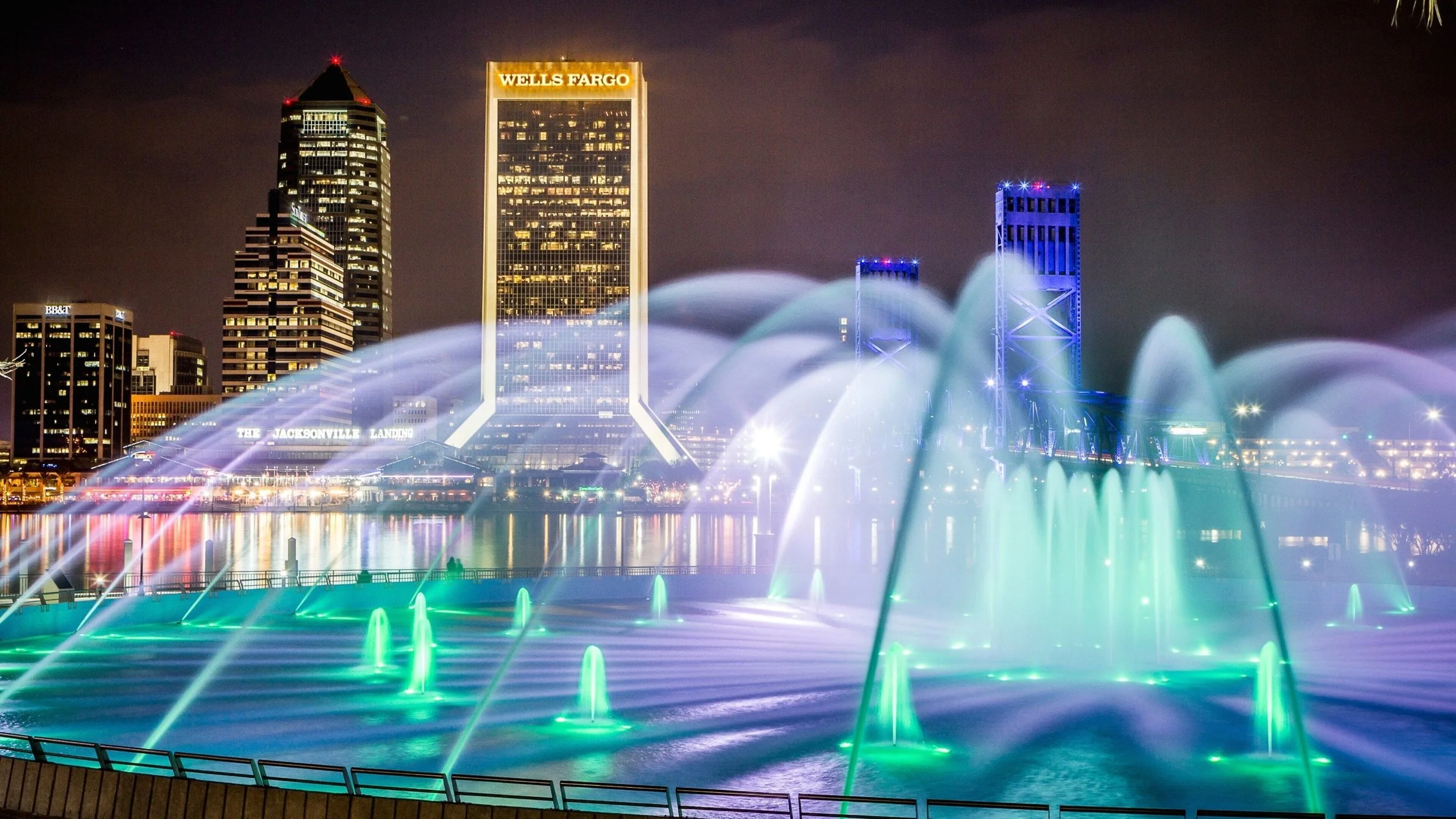 Nighttime view of downtown Jacksonville with illuminated skyscrapers and a water fountain display with colorful lights.