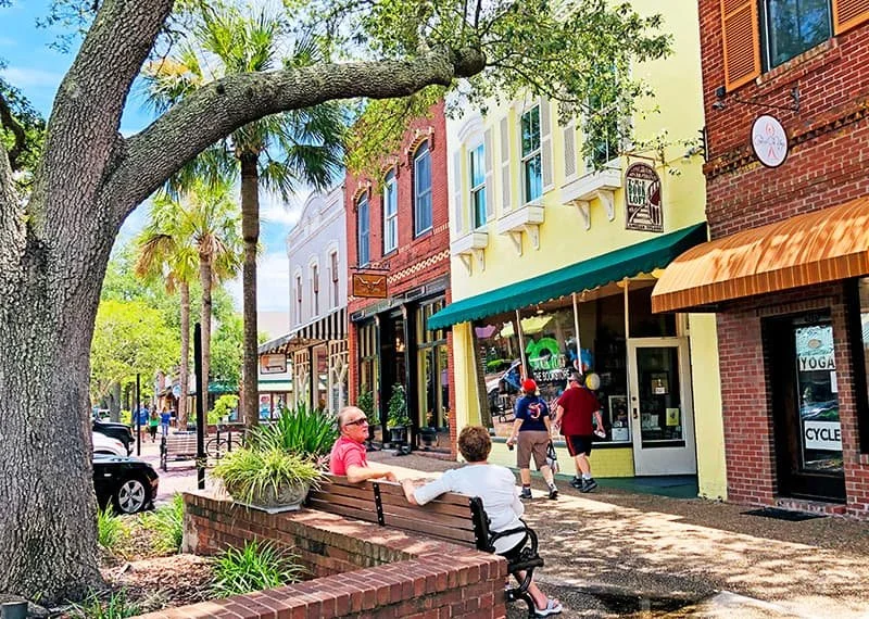 People sitting on a bench under a large tree in front of a row of small shops with colorful facades on a sunny day.